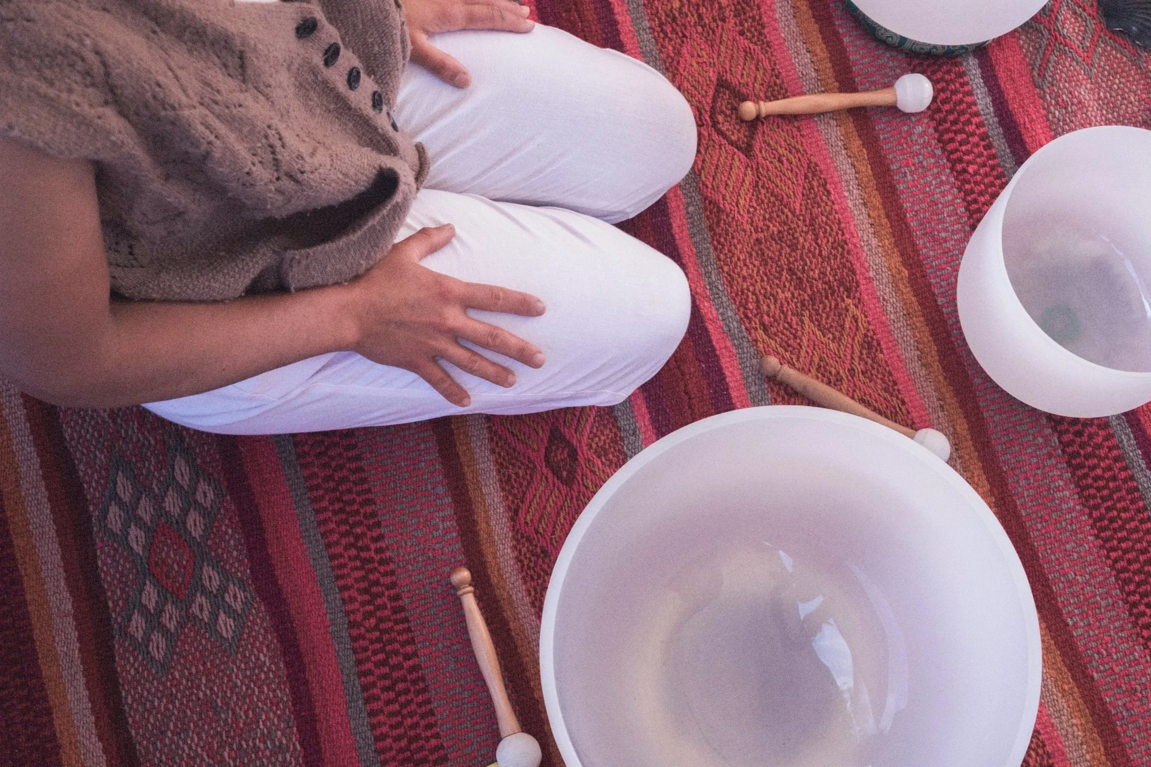Person kneeling on a colorful patterned rug, wearing white pants and a brown sweater, with parts of white bowls and wooden pegs visible on the rug.