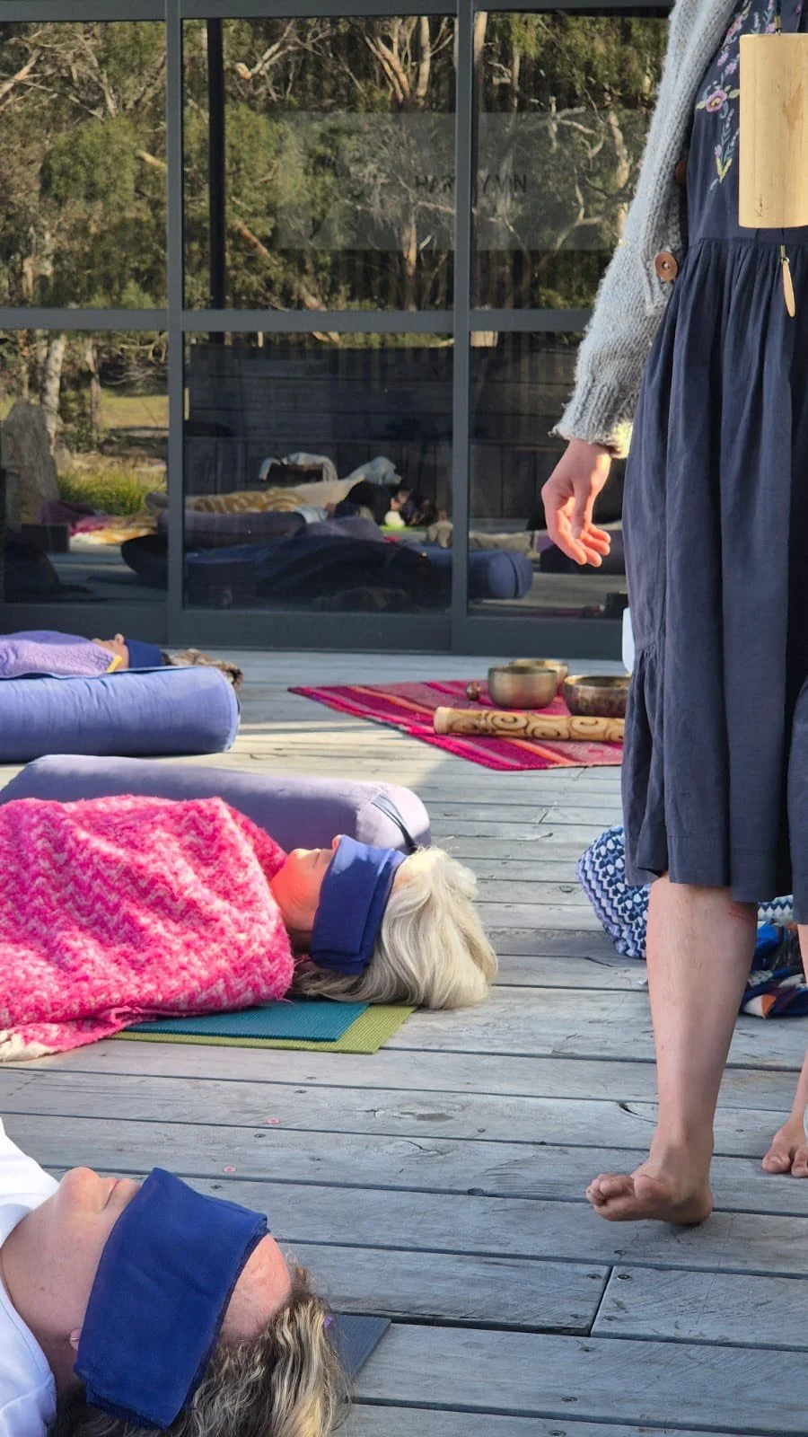 People participating in a sound bath and breath work practice outdoors on a wooden deck, lying on mats with eyes covered, while immersed in the sound of wind chimes.