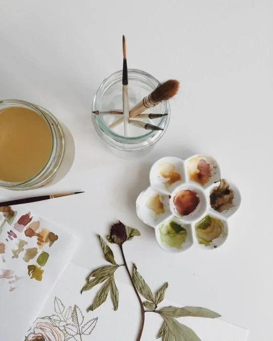 Paint palette with various colors, paintbrushes in a jar, a paintbrush on a white surface, and botanical illustrations on paper.