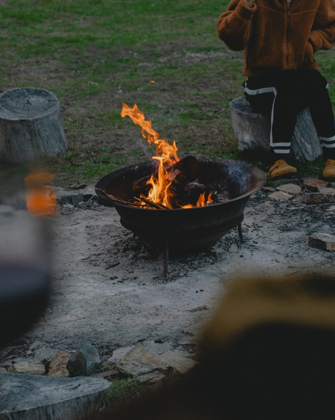 A fire pit with flames burning outdoors, surrounded by logs and people sitting nearby.