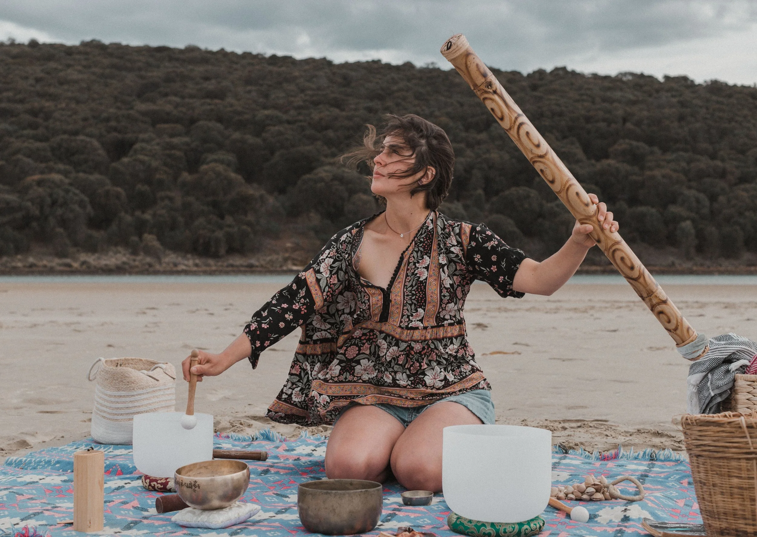 Woman with short hair, wearing a floral patterned dress, sitting cross-legged on a blanket on sandy beach, holding a wooden baton, surrounded by bowls and a basket with stones, with a hilly landscape and cloudy sky in the background.