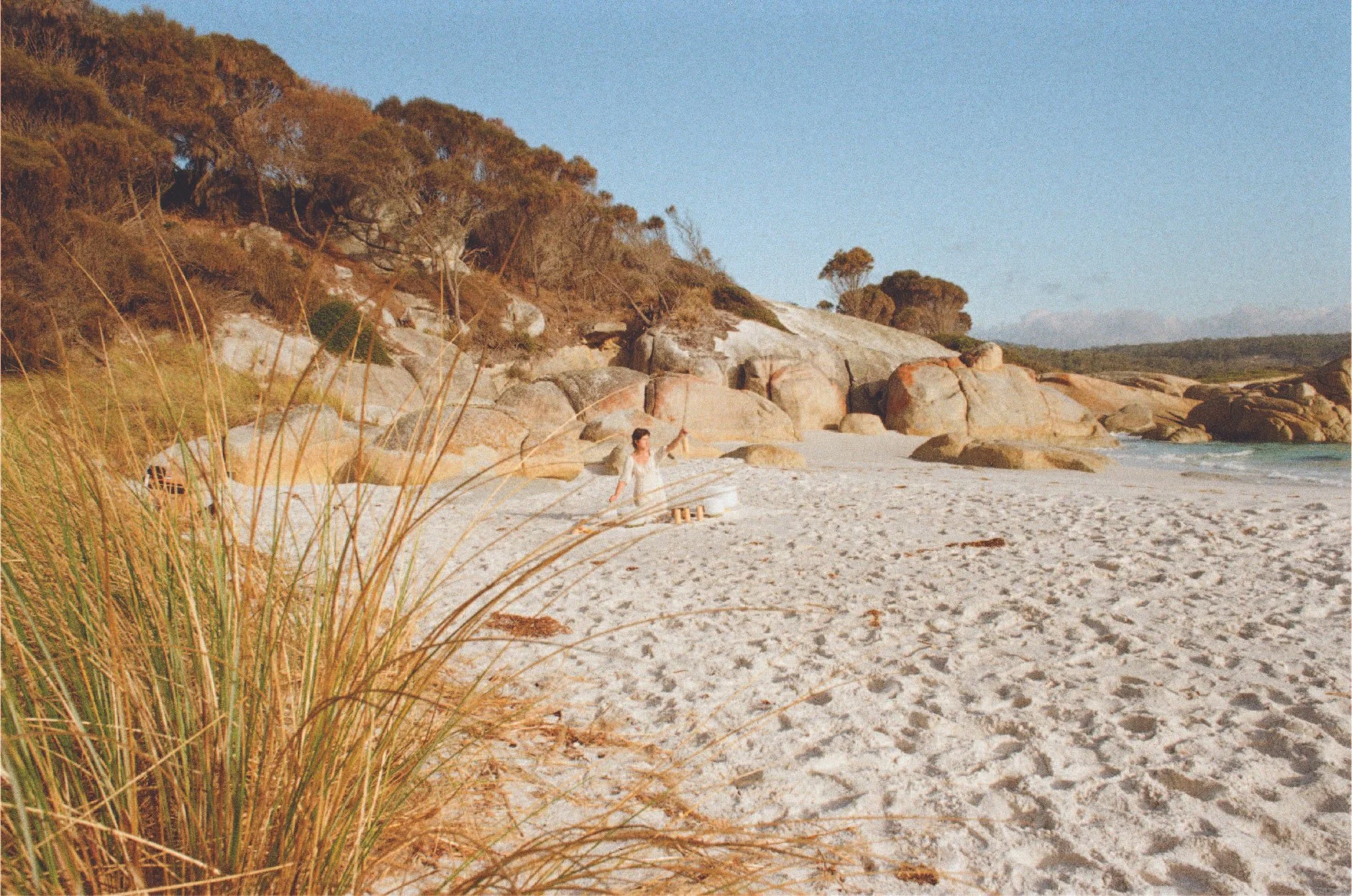 A person in white clothing standing on a sandy beach near large rocks and trees, with dunes and vegetation in the foreground and a clear sky in the background.