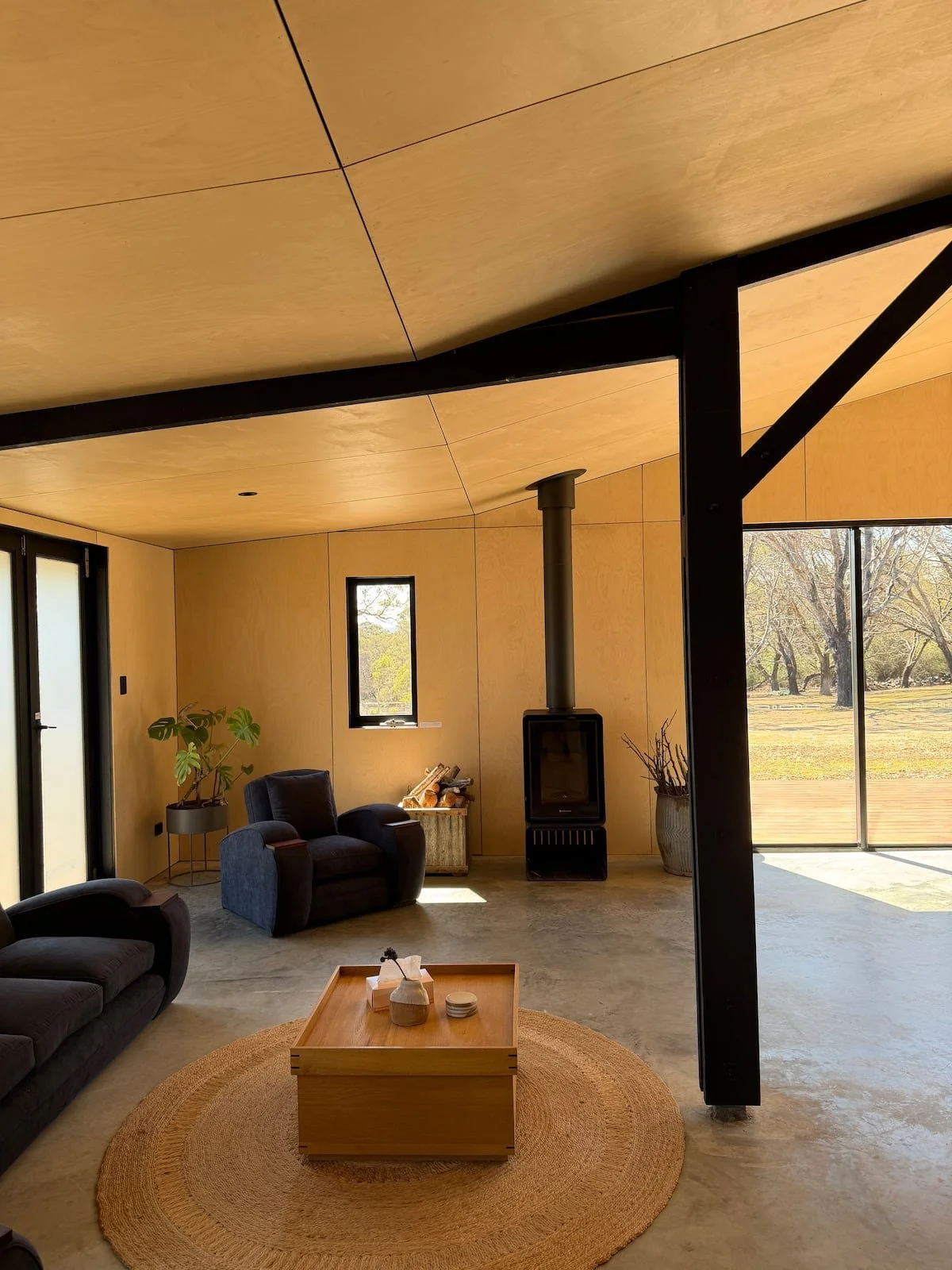 Modern living room with wooden walls and ceiling, black sofas, a round woven rug, a wooden coffee table, a wood stove, and large windows showing outdoor trees.