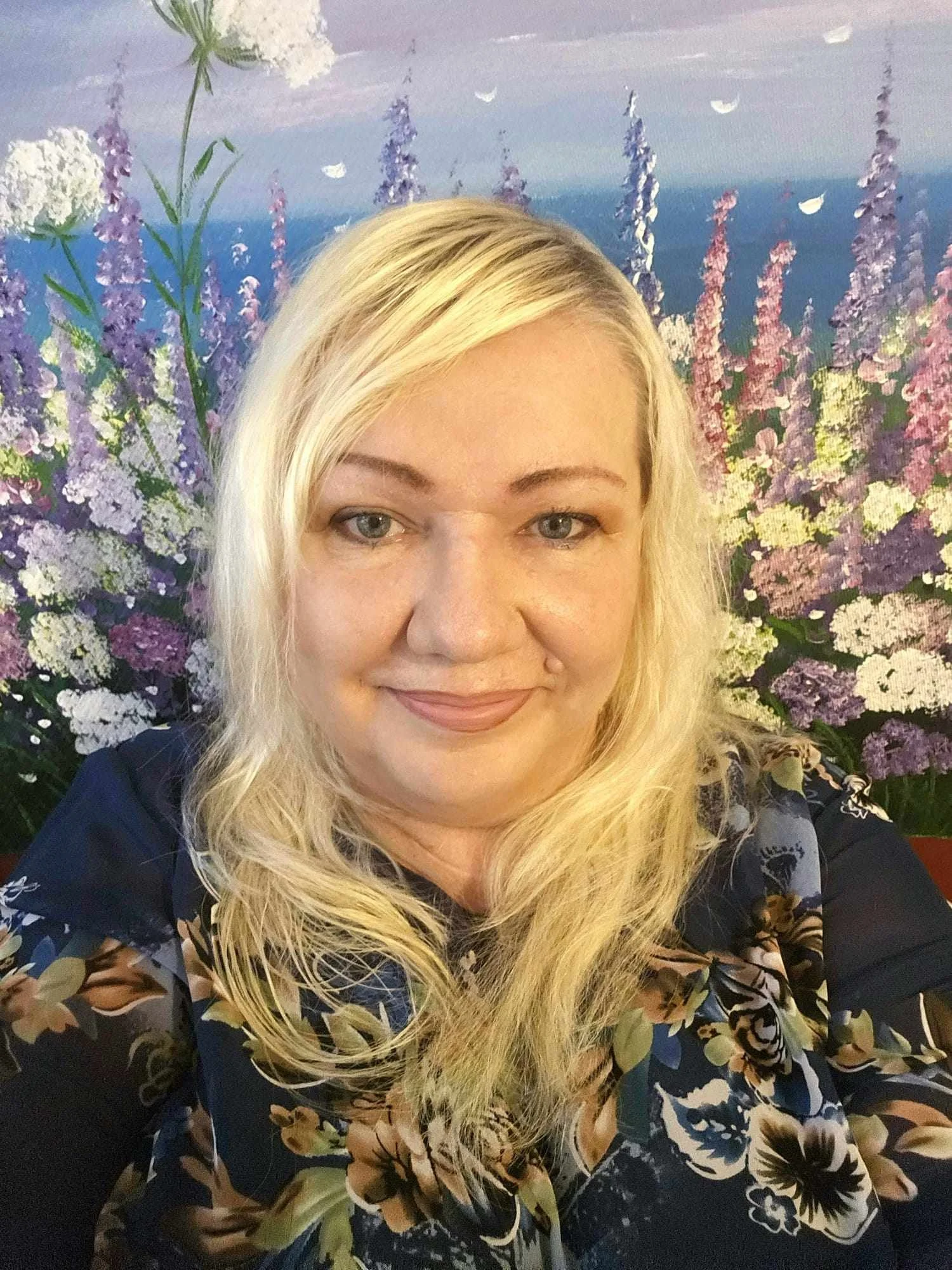 A woman with long blonde hair and blue eyes taking a selfie in front of a floral mural with purple, white, and pink flowers and a blue sky background.