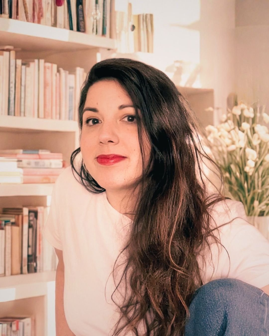 A woman with long, dark hair and red lipstick taking a selfie in a room with bookshelves and white flowers in the background.
