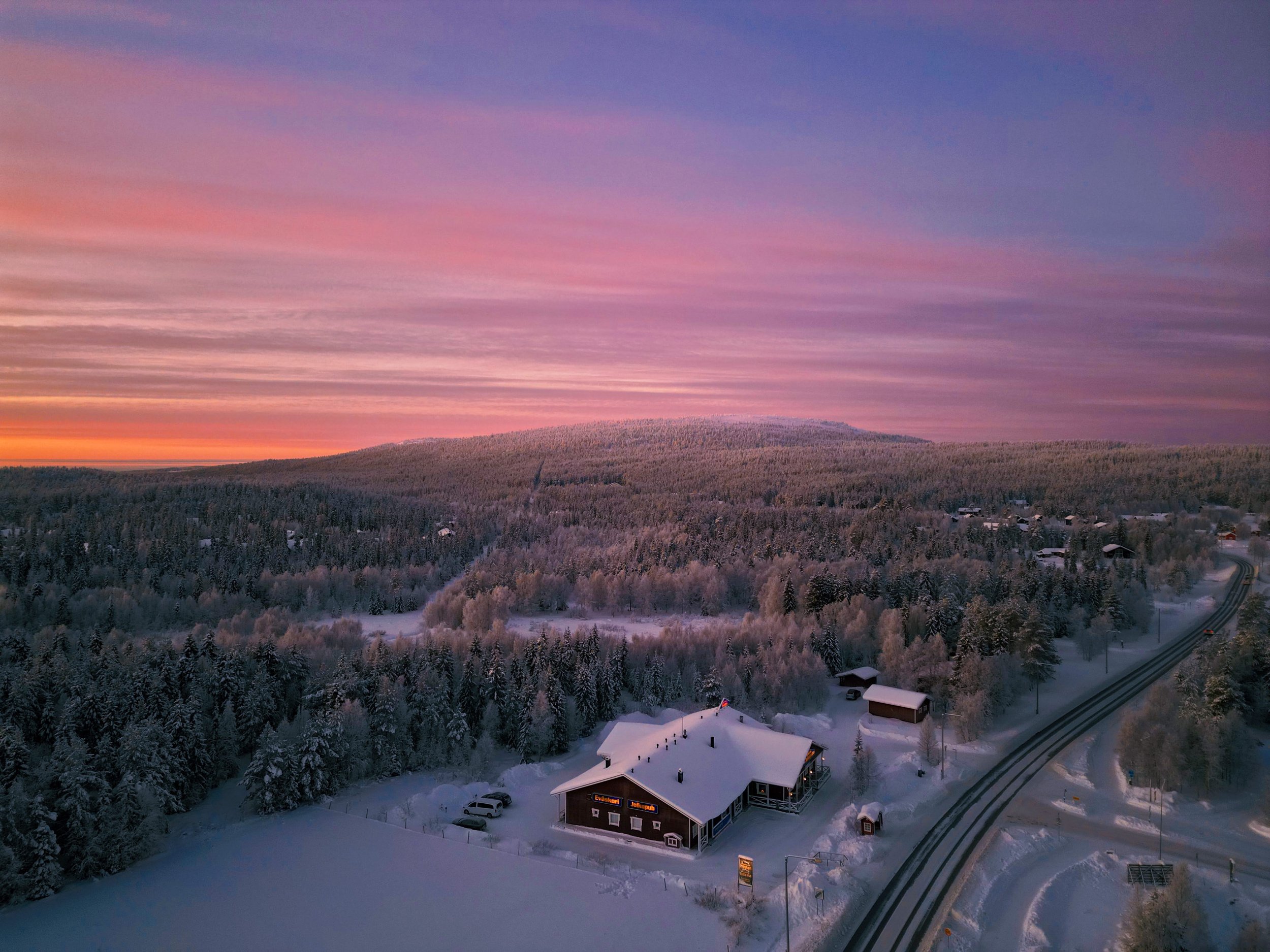 Talvi-ilta Ylläs-tunturilla, jossa on luminen metsä ja rakennuksia, joista on valaistus. Taivaalla on pilviä ja sävyjä auringonlaskusta, kuten vaaleanpunaista, lilaa ja oranssia.