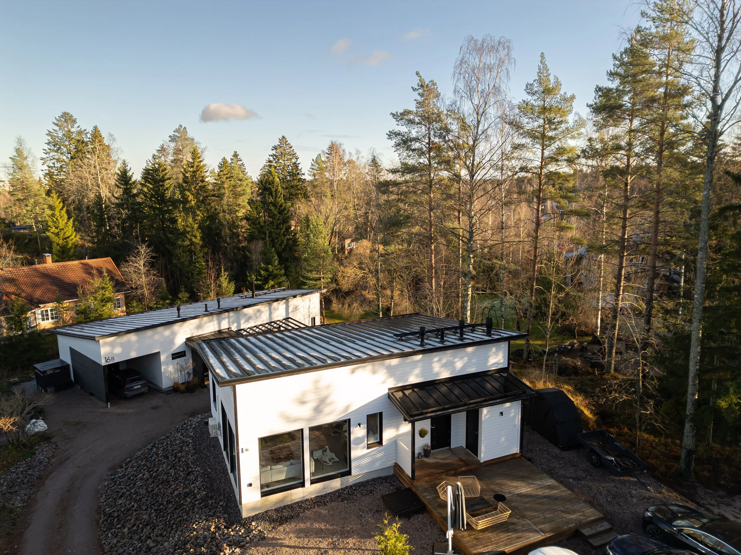 Moderni valkoinen talo, ympäröi metsäiset alueet ja muita rakennuksia, kova katto ja iso lasinen etuosa näkyvät.