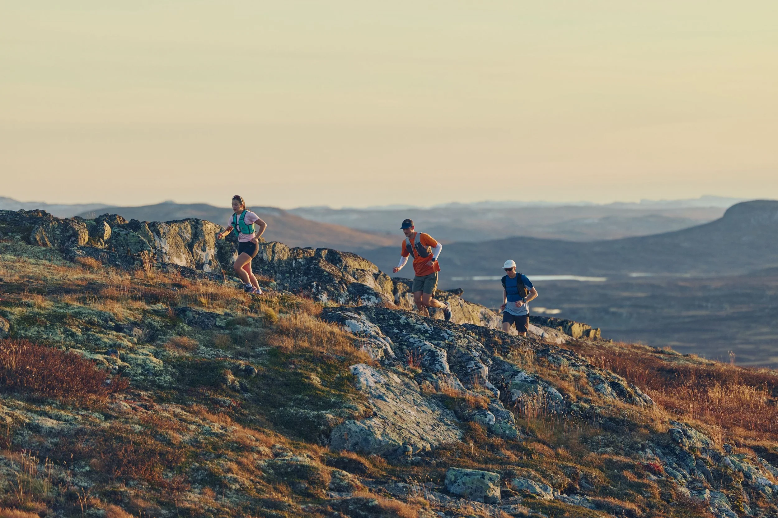 Three hikers wearing backpacks trekking on a rocky hillside during sunset.