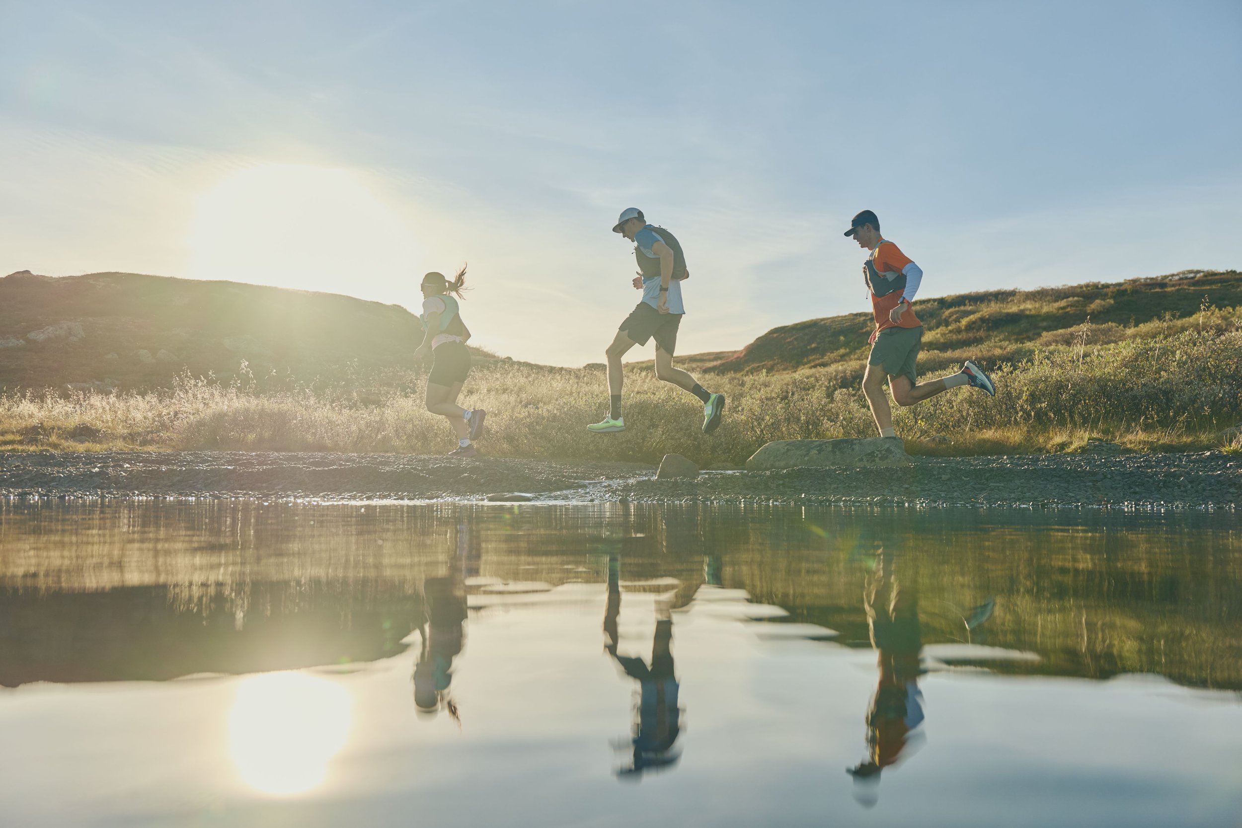 Four children are jumping into a body of water during sunset on a grassy landscape.