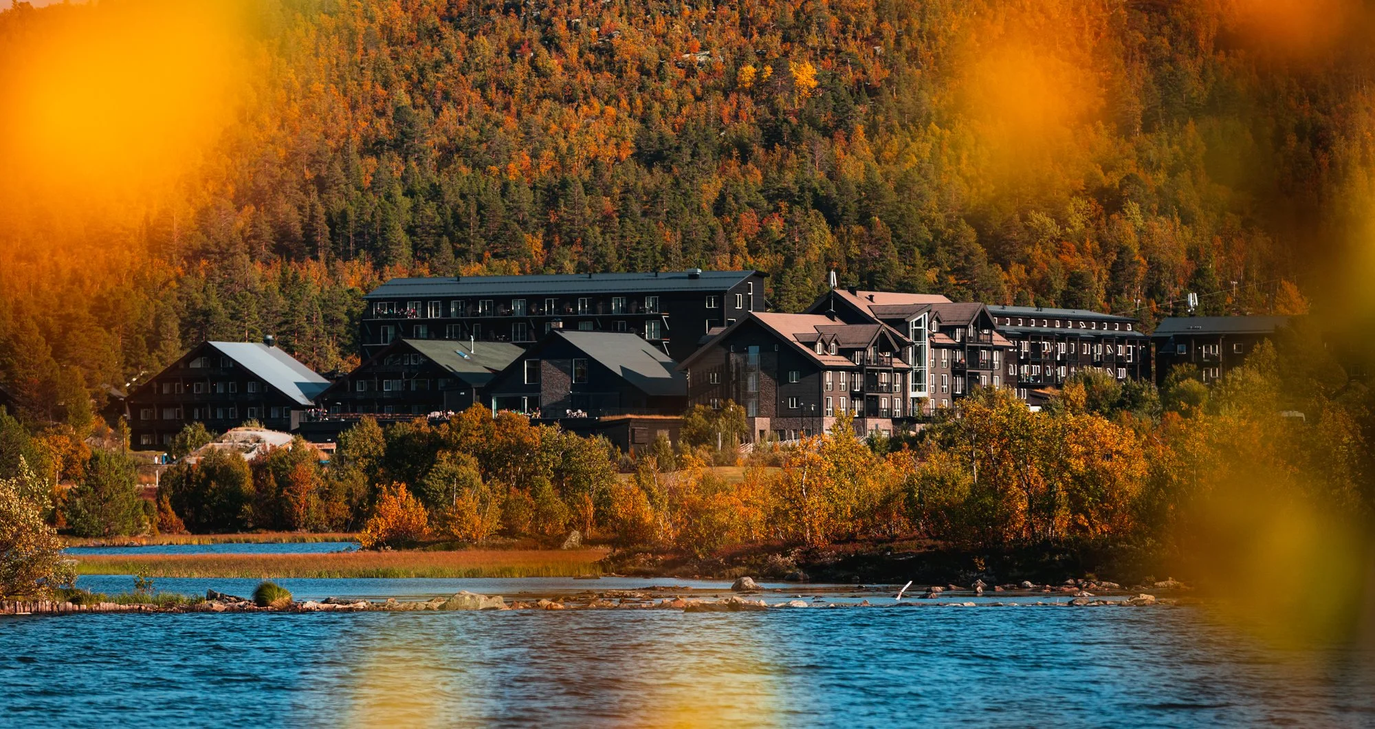 A lakeside view of a resort with black buildings surrounded by autumn foliage, with a forested mountain in the background.