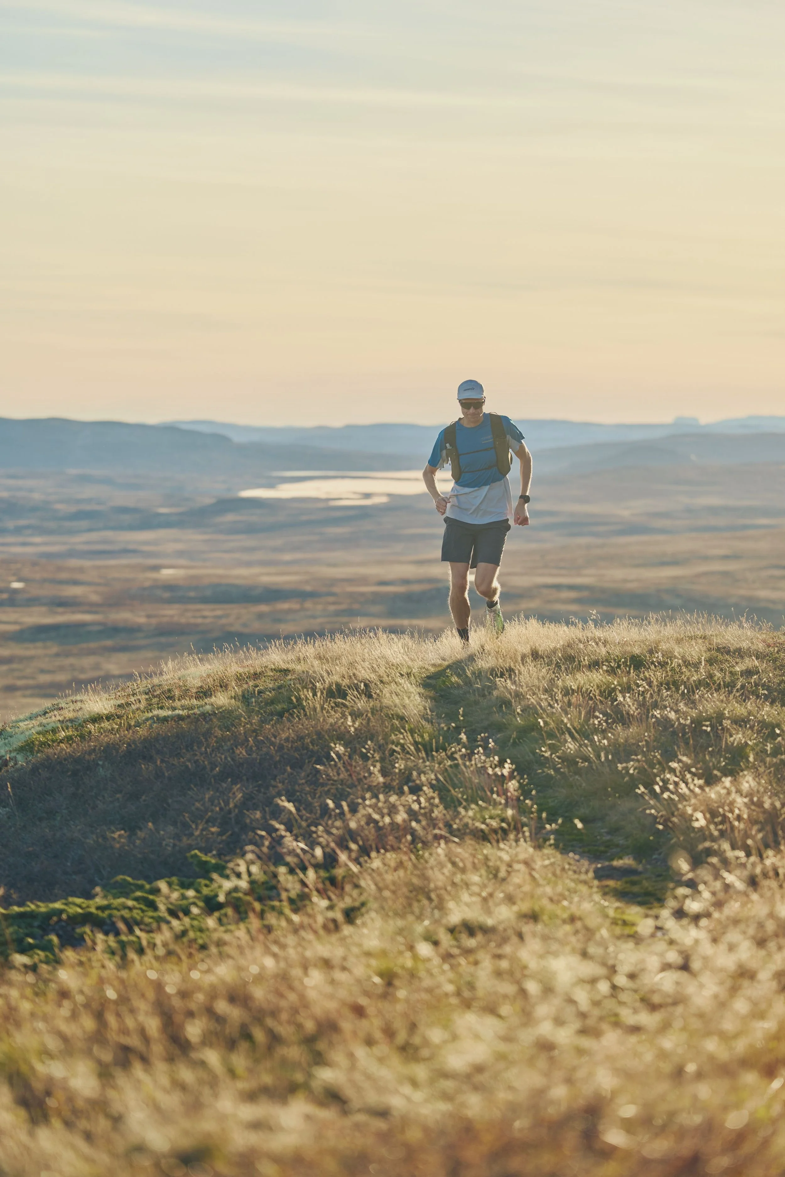 A person trail running on a grassy hill during sunset or sunrise, wearing a blue t-shirt, black shorts, a white cap, sunglasses, and a backpack.