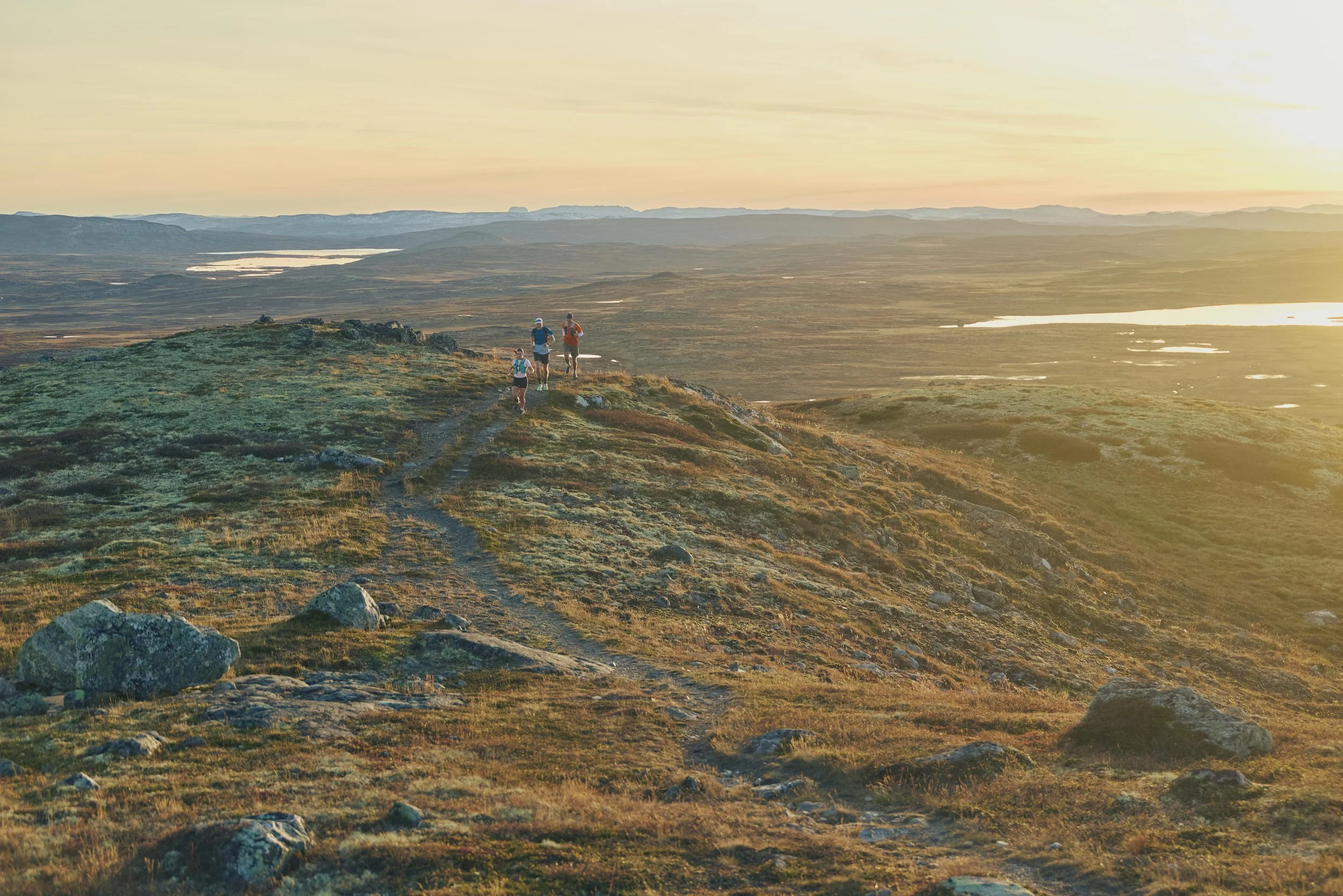 Four hikers walking along a trail on a grassy, rocky hill during sunset in a wide open landscape with lakes in the distance.