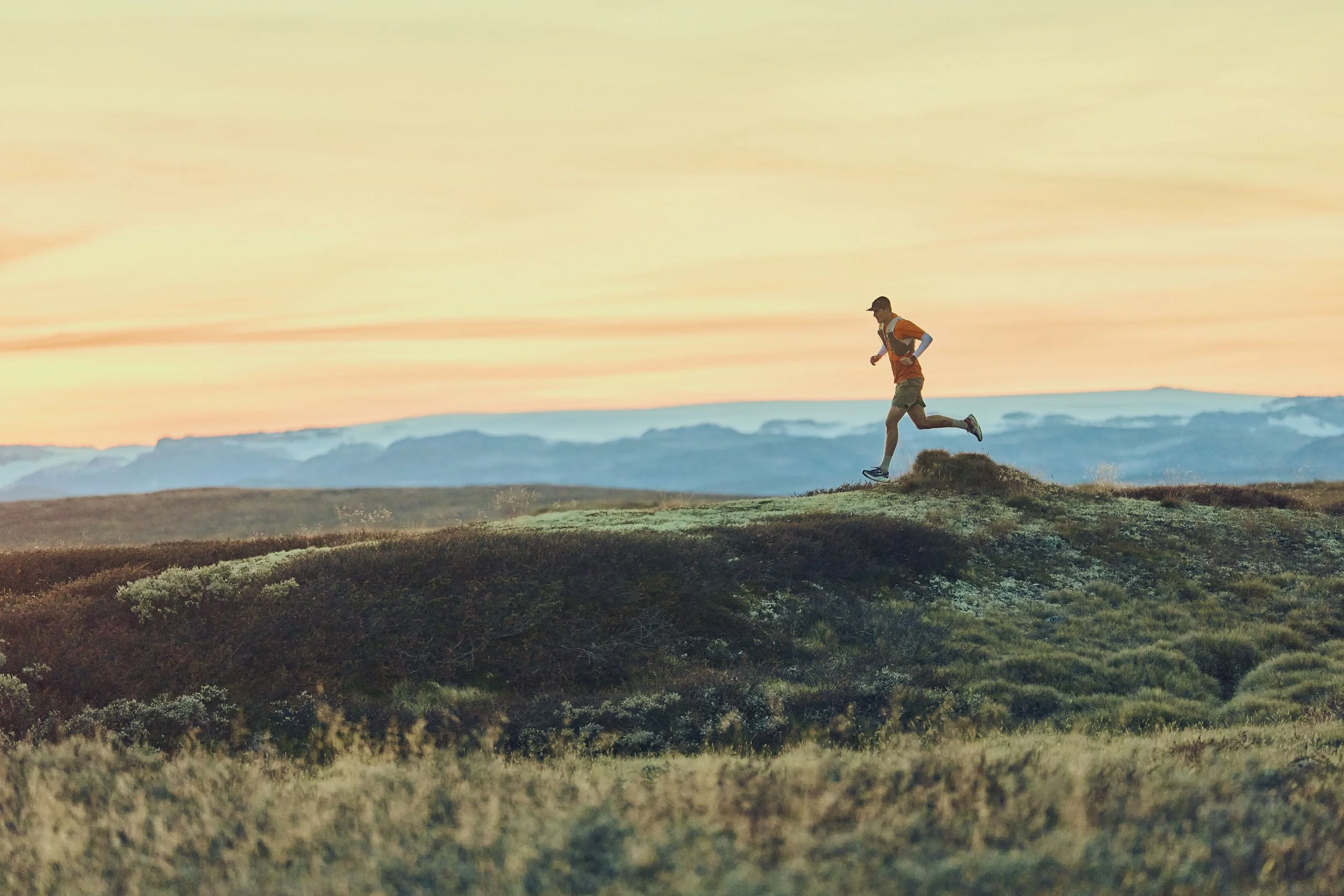 A person running on a grassy hill at sunset with mountains in the background.