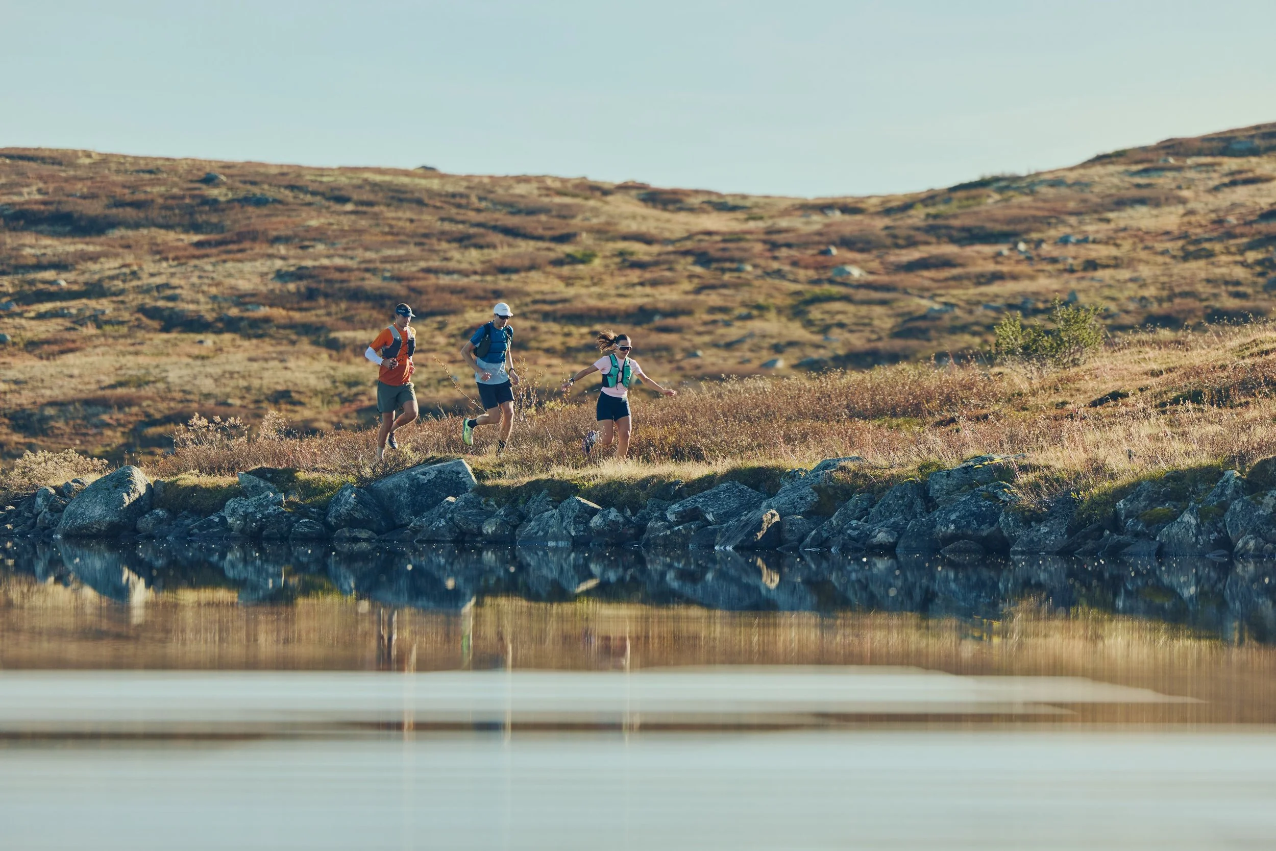 Three hikers walking along a rocky shoreline beside calm water in a mountainous area with dry grass and sparse vegetation.