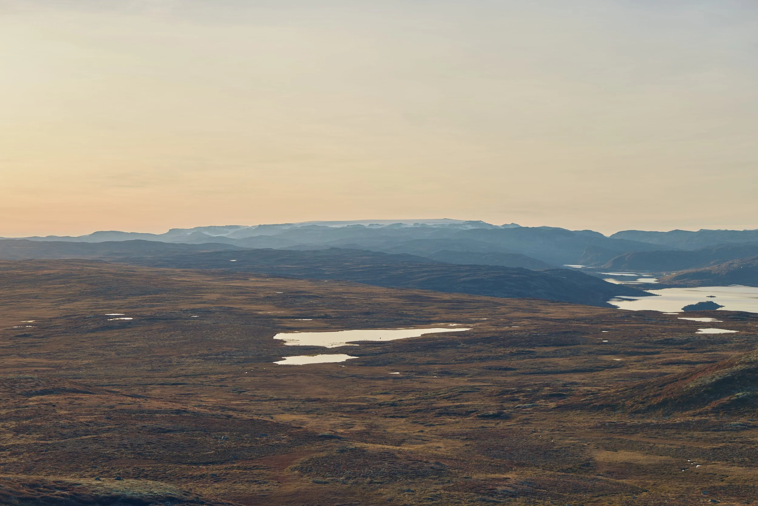 A vast, open landscape with rolling hills, small lakes, and distant mountain ranges under a pale, cloudy sky.