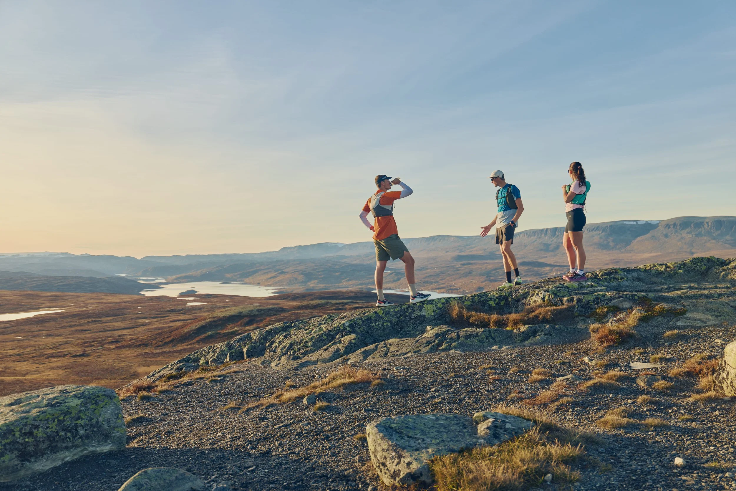 Three hikers, two men and one woman, standing on a rocky landscape in a mountainous area with a river in the distance during sunset.