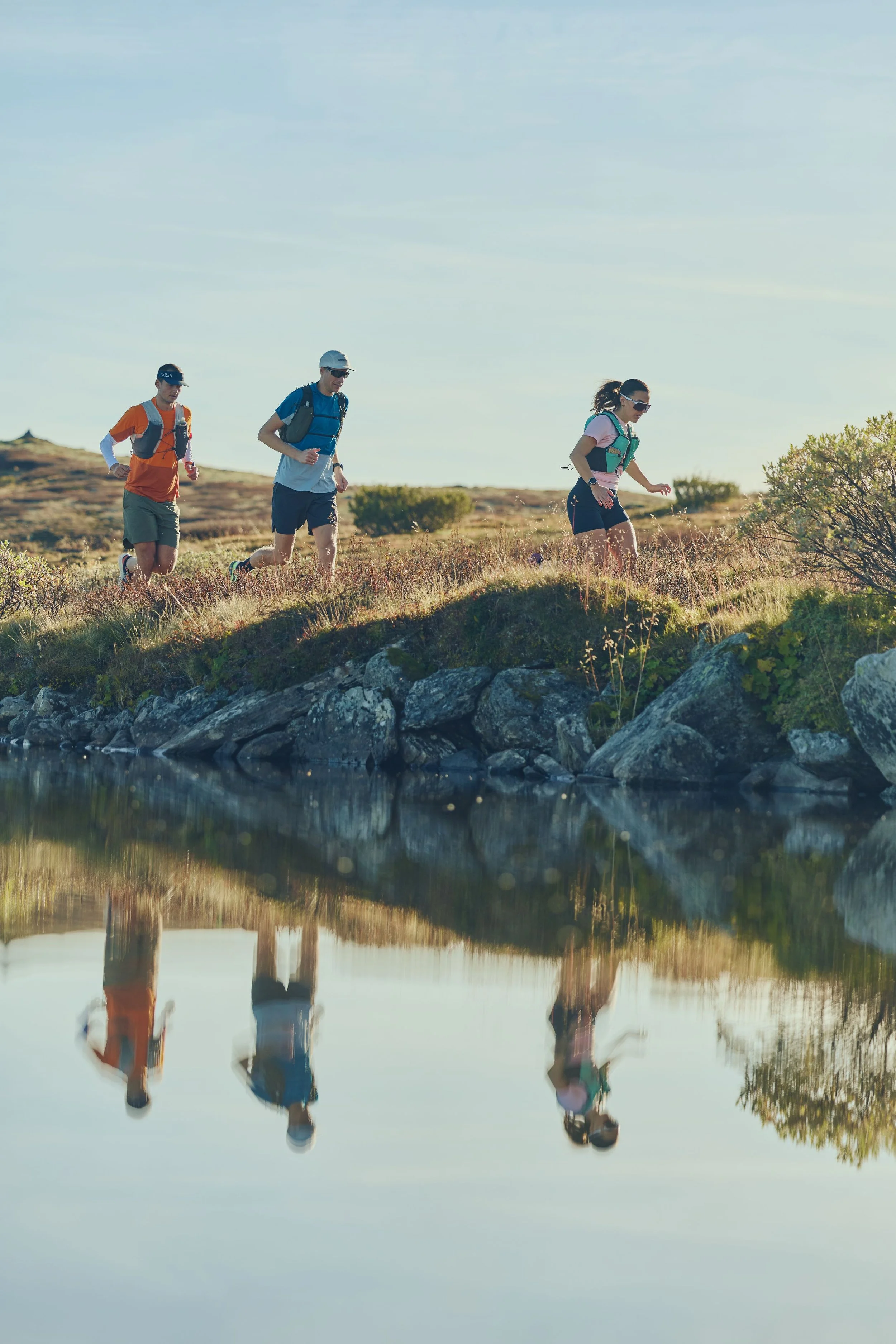Three people trail running along a rocky bank by a calm water body in a natural outdoor setting with grassy terrain and sparse shrubs.