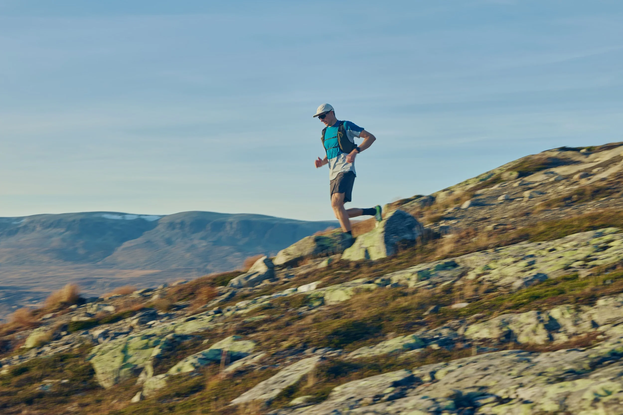 A person trail running on a rocky hillside with mountains in the background under a clear sky.