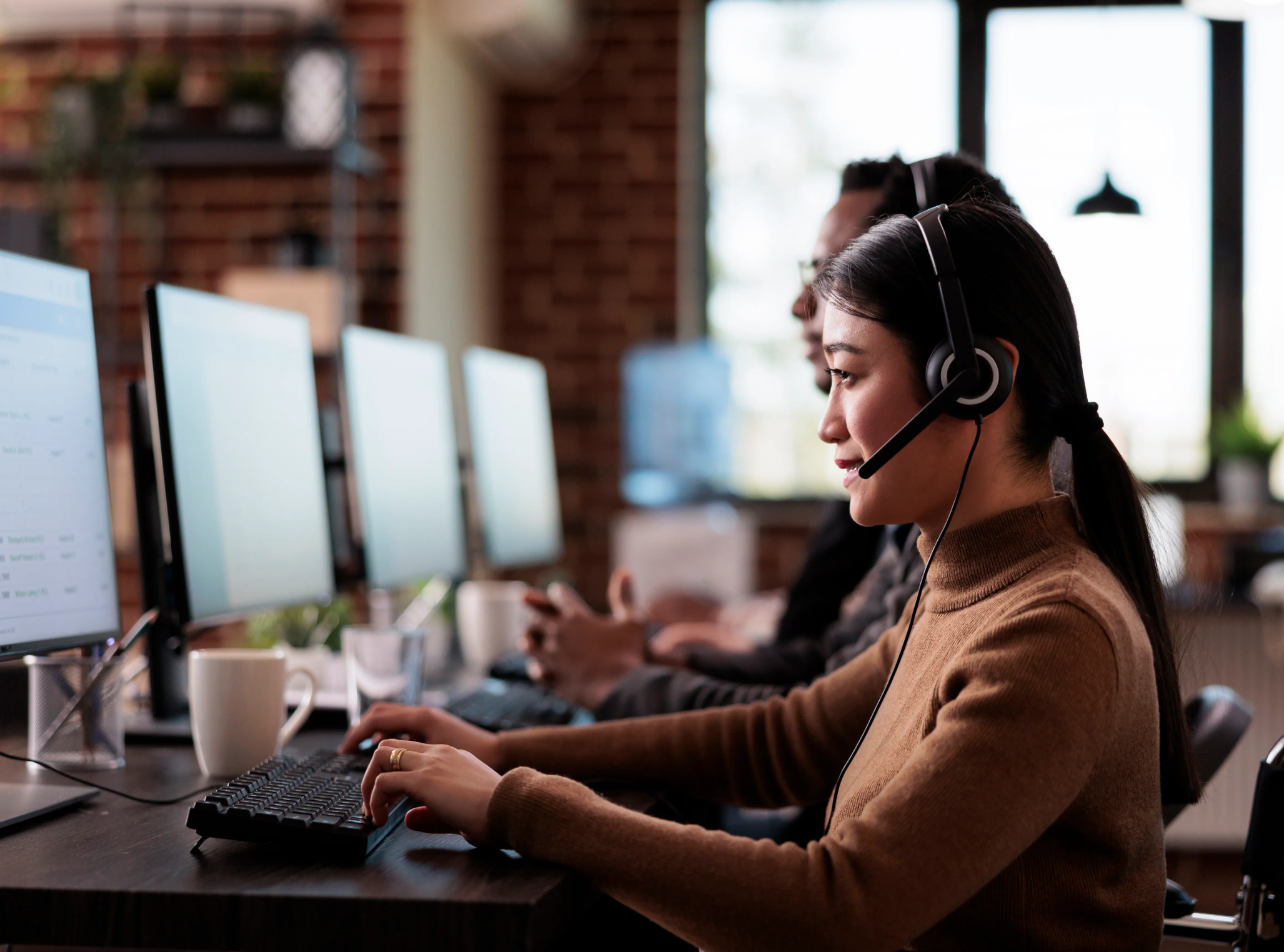 A woman wearing a headset working at a computer in an office with colleagues.