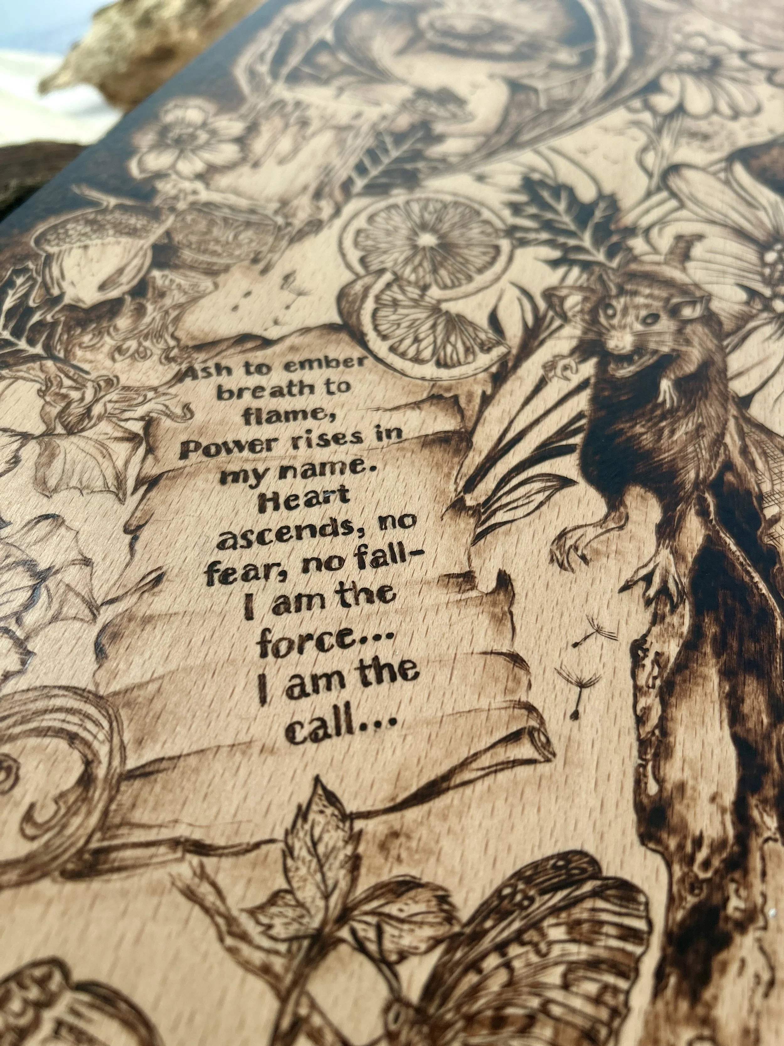 Close-up of a hand-lettered poem on a scroll, featuring the text "Ash to ember / breath to flame / Power rises in my name," burned into wood.