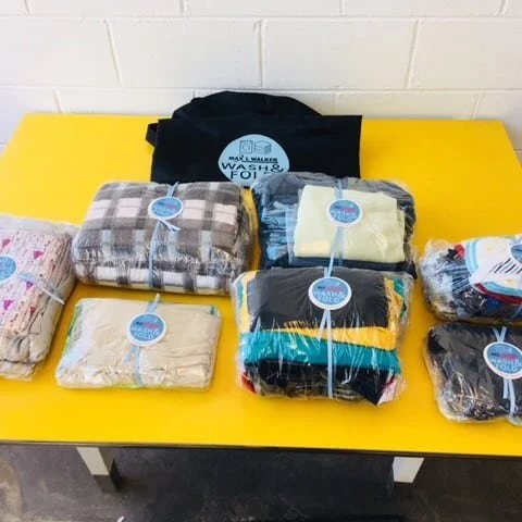 Multiple folded blankets wrapped in plastic on a yellow table, with a black reusable bag in the background.