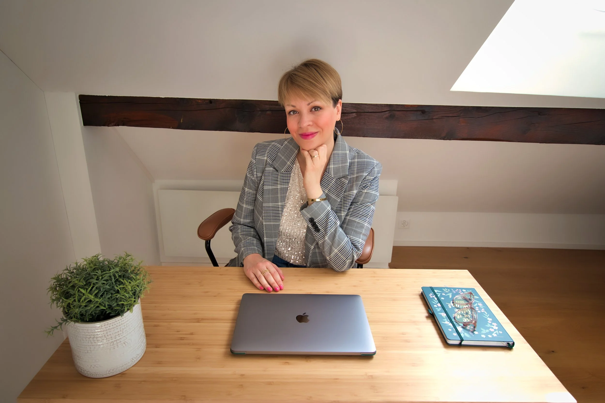 Woman sitting at desk under sloped ceiling, smiling, with laptop, glasses, notebook, and houseplant.