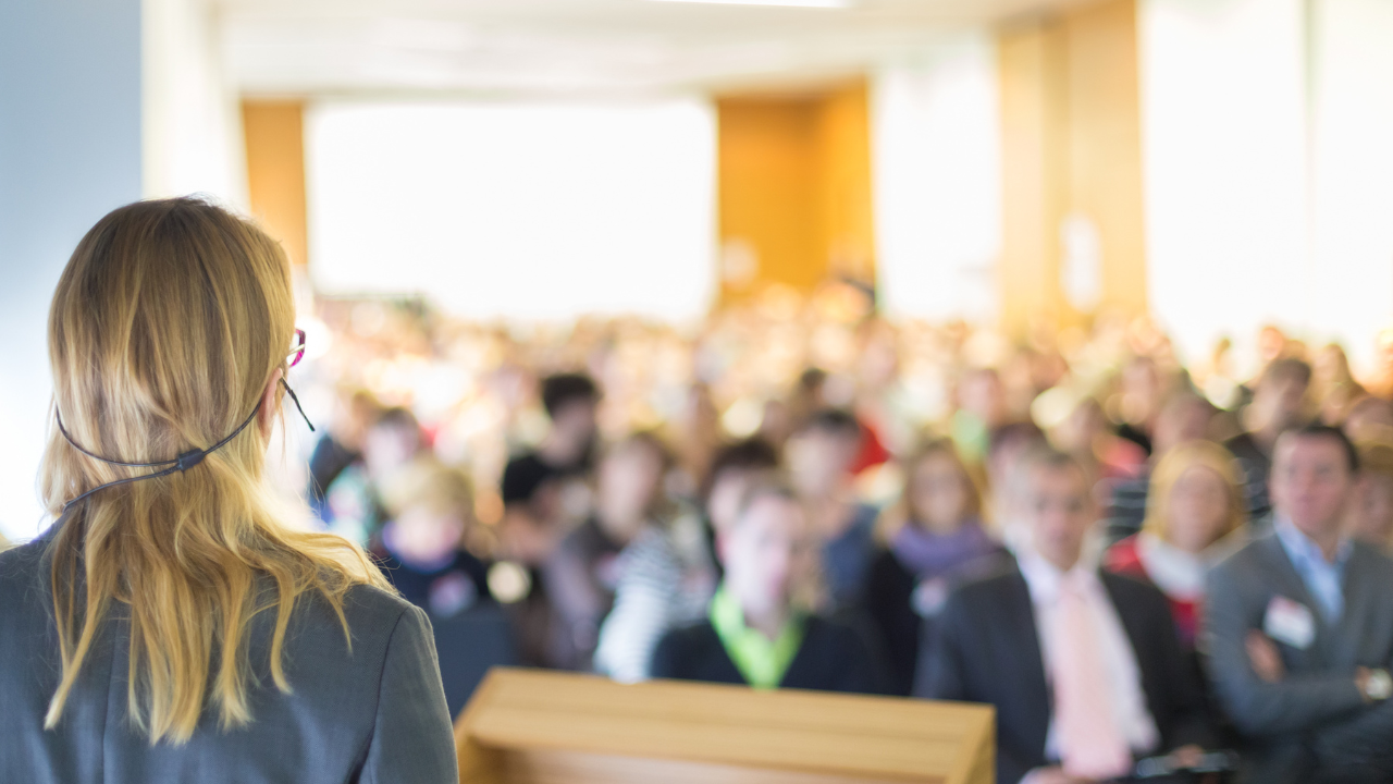 A woman with red hair and glasses giving a presentation to a large audience in a conference room.