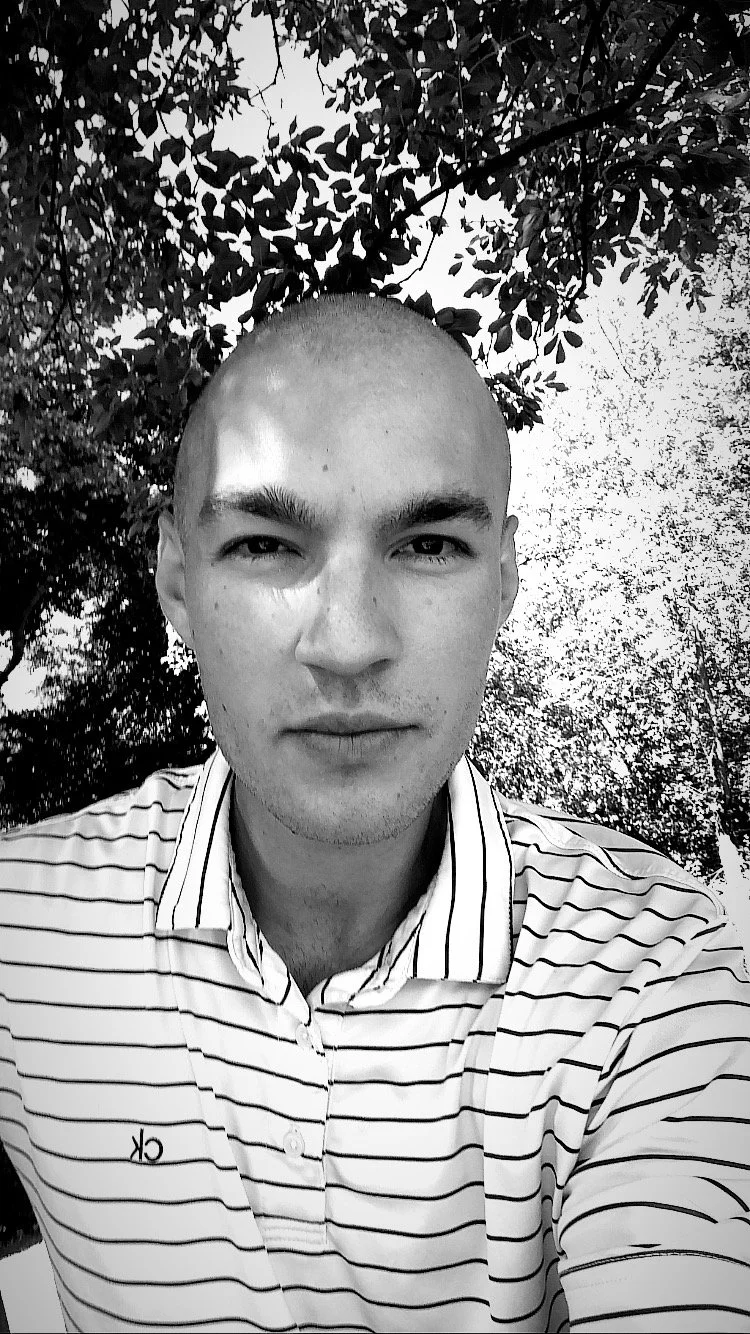 Schwarz-weiße Nahaufnahme eines jungen Mannes mit kurzem Haar, der vor Bäumen und Blättern steht und direkt in die Kamera schaut.