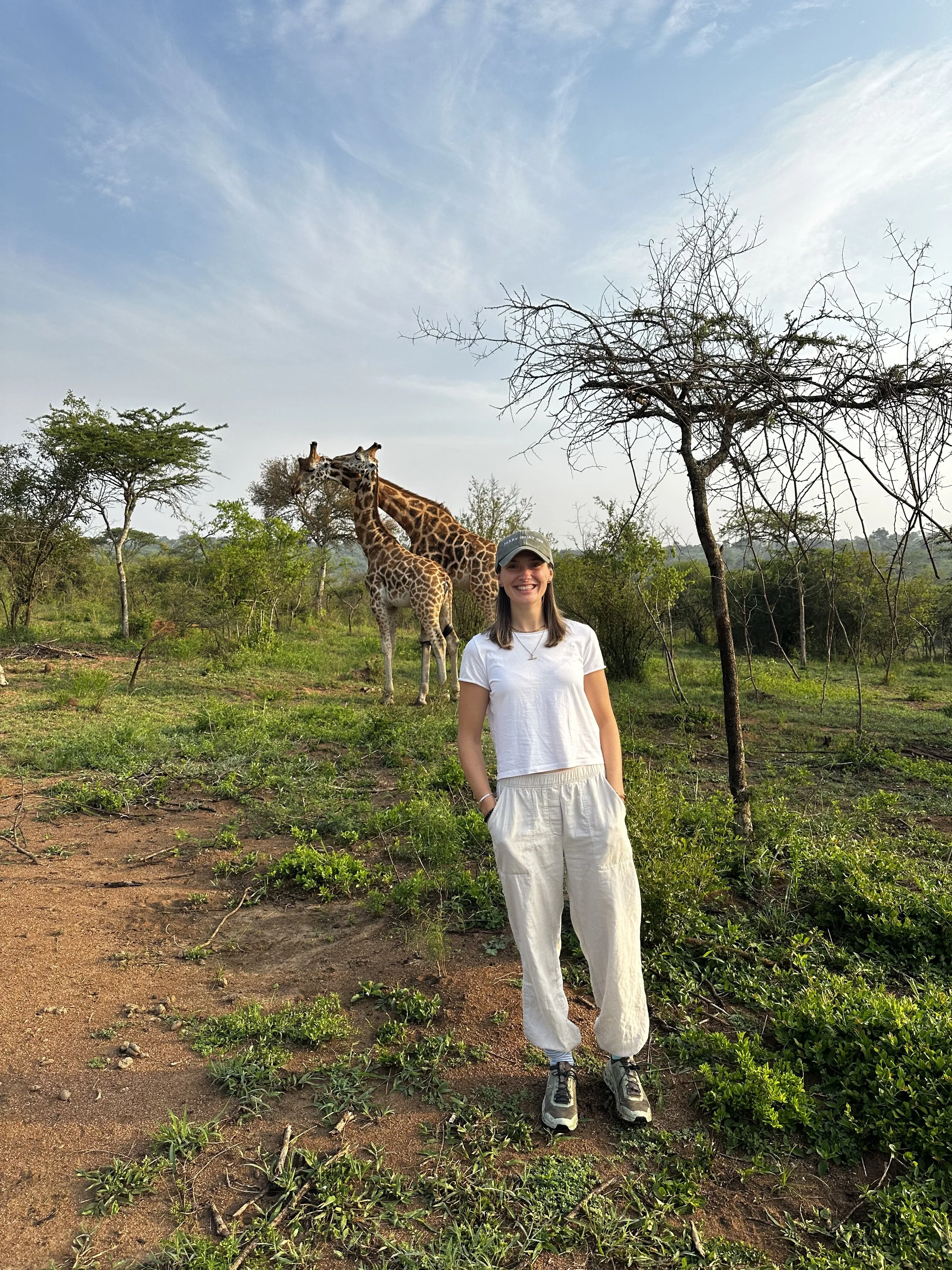 A woman smiling and standing outdoors in a savannah landscape with two giraffes in the background.