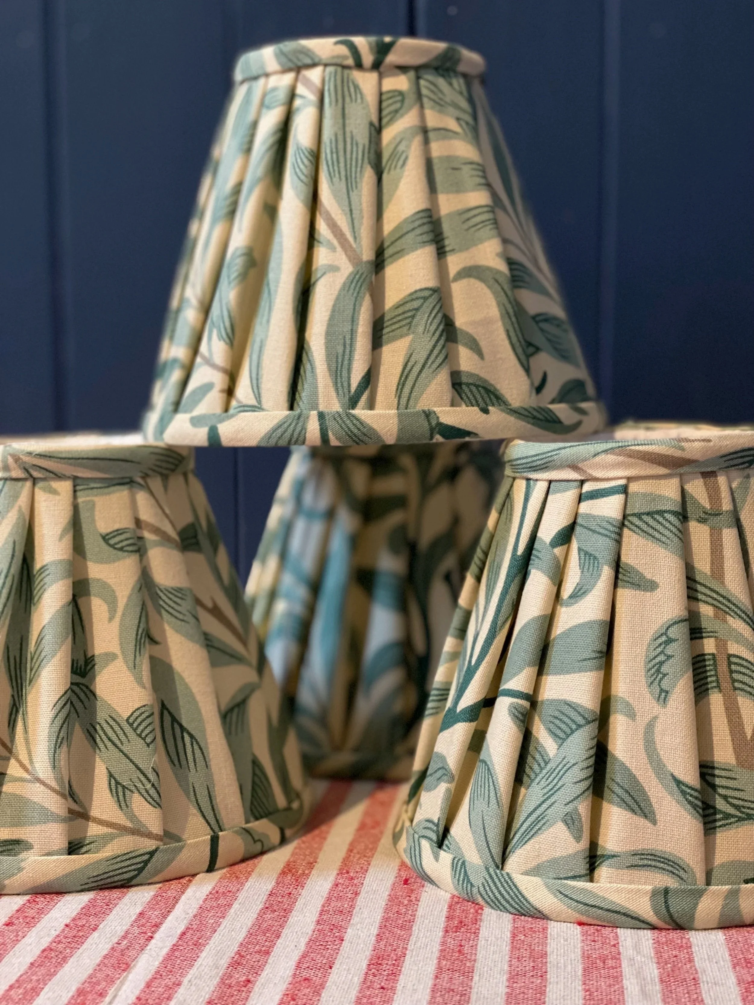 Three fabric lampshades with a green leaf pattern on a pink and white striped tablecloth against a blue backdrop.
