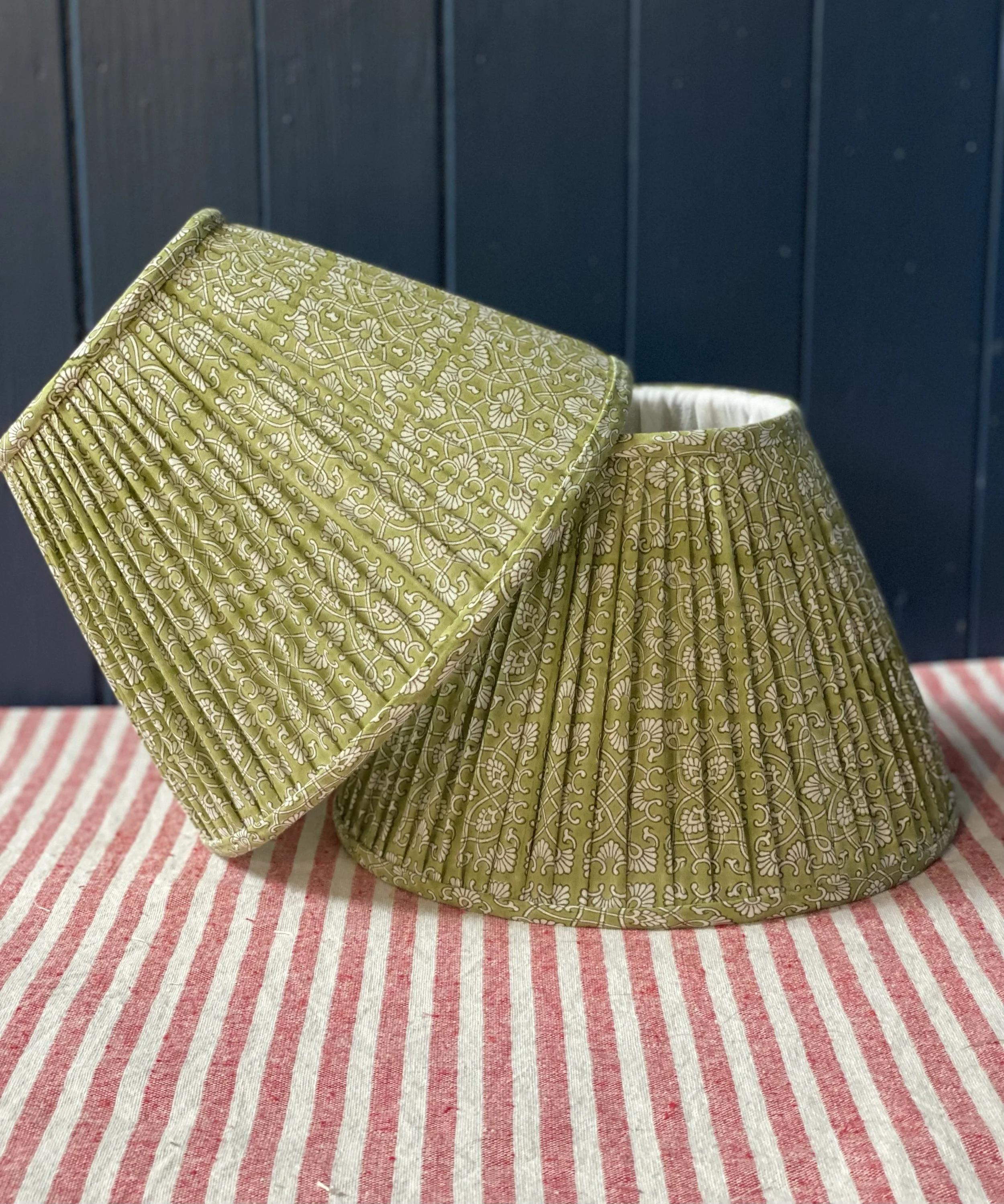 A folded decorative lampshade with intricate pattern in green and white, placed on a red and white striped cloth with a blue wall background.