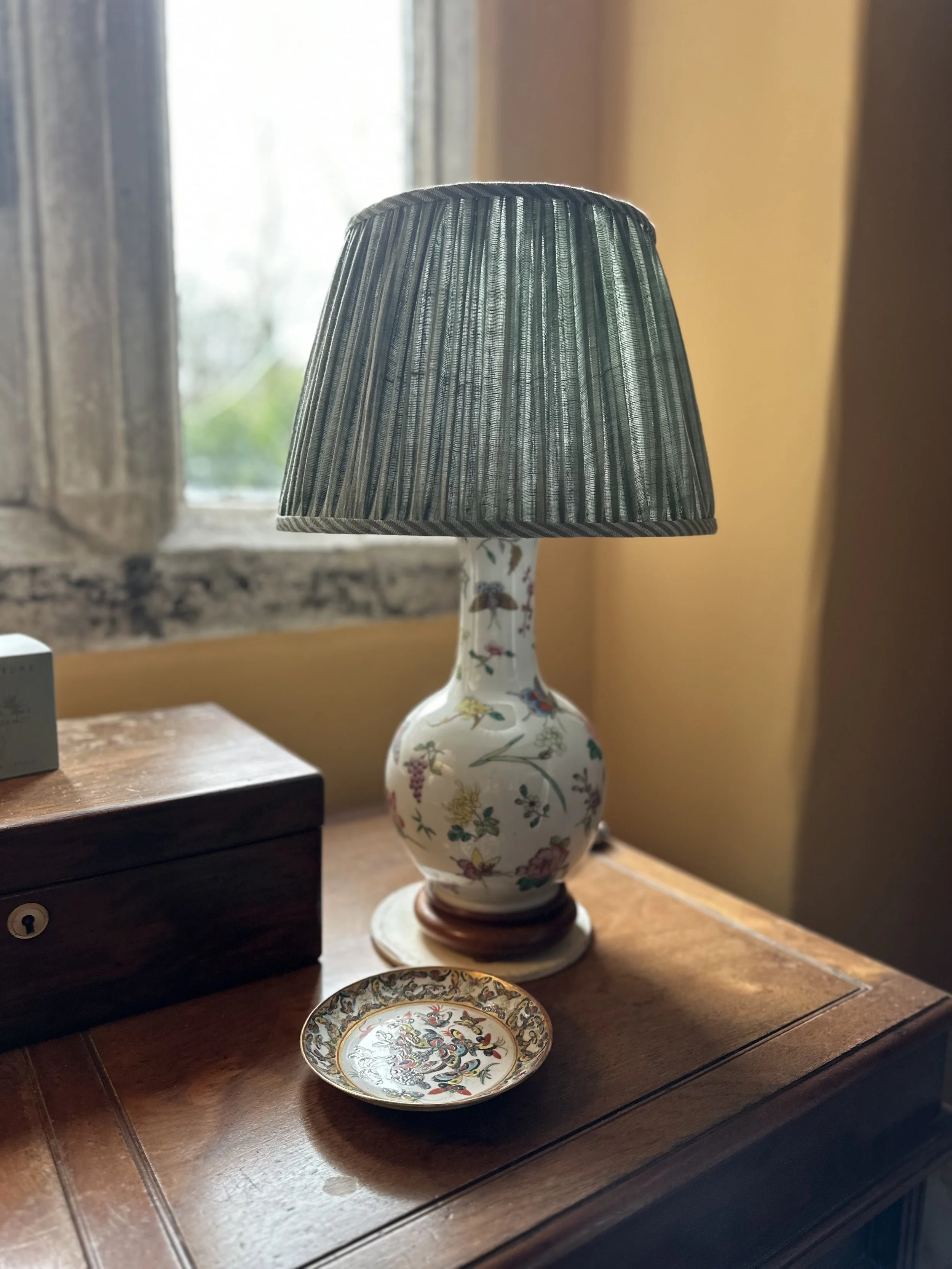 A floral-patterned ceramic table lamp with a dark green pleated shade on a wooden surface. A small decorative dish with intricate patterns is in front of it, and a window with a stone sill is in the background.