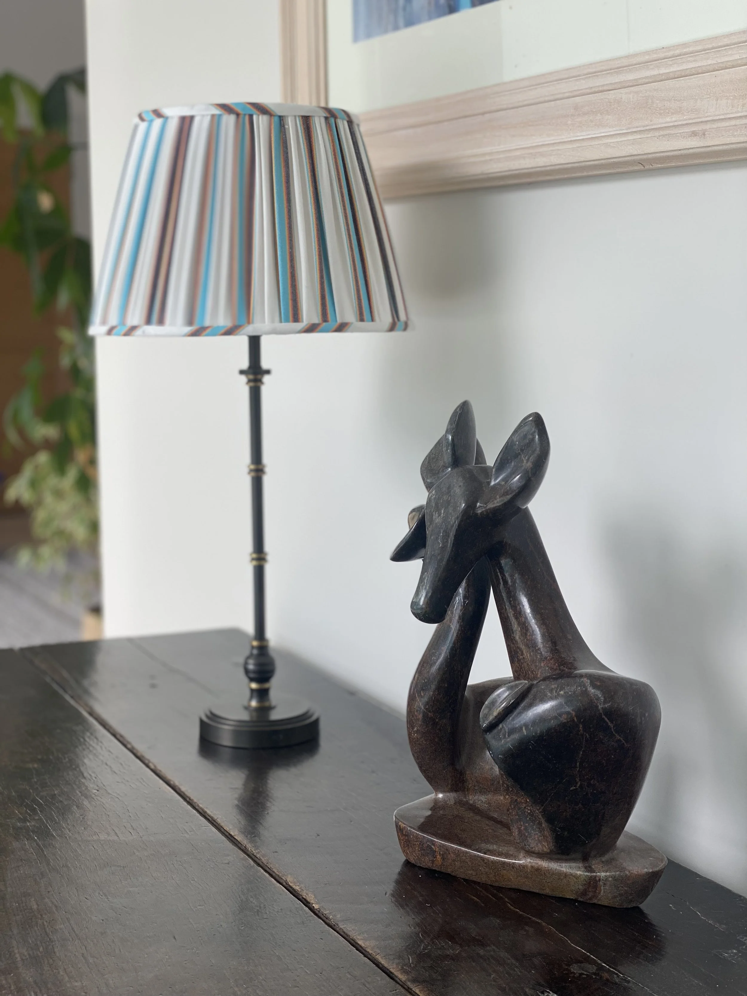 A decorative sculpture of a sitting animal, likely a dog, made of dark marble or stone, placed on a wooden surface next to a table lamp with a colorful striped shade, against a white wall with a framed picture above.