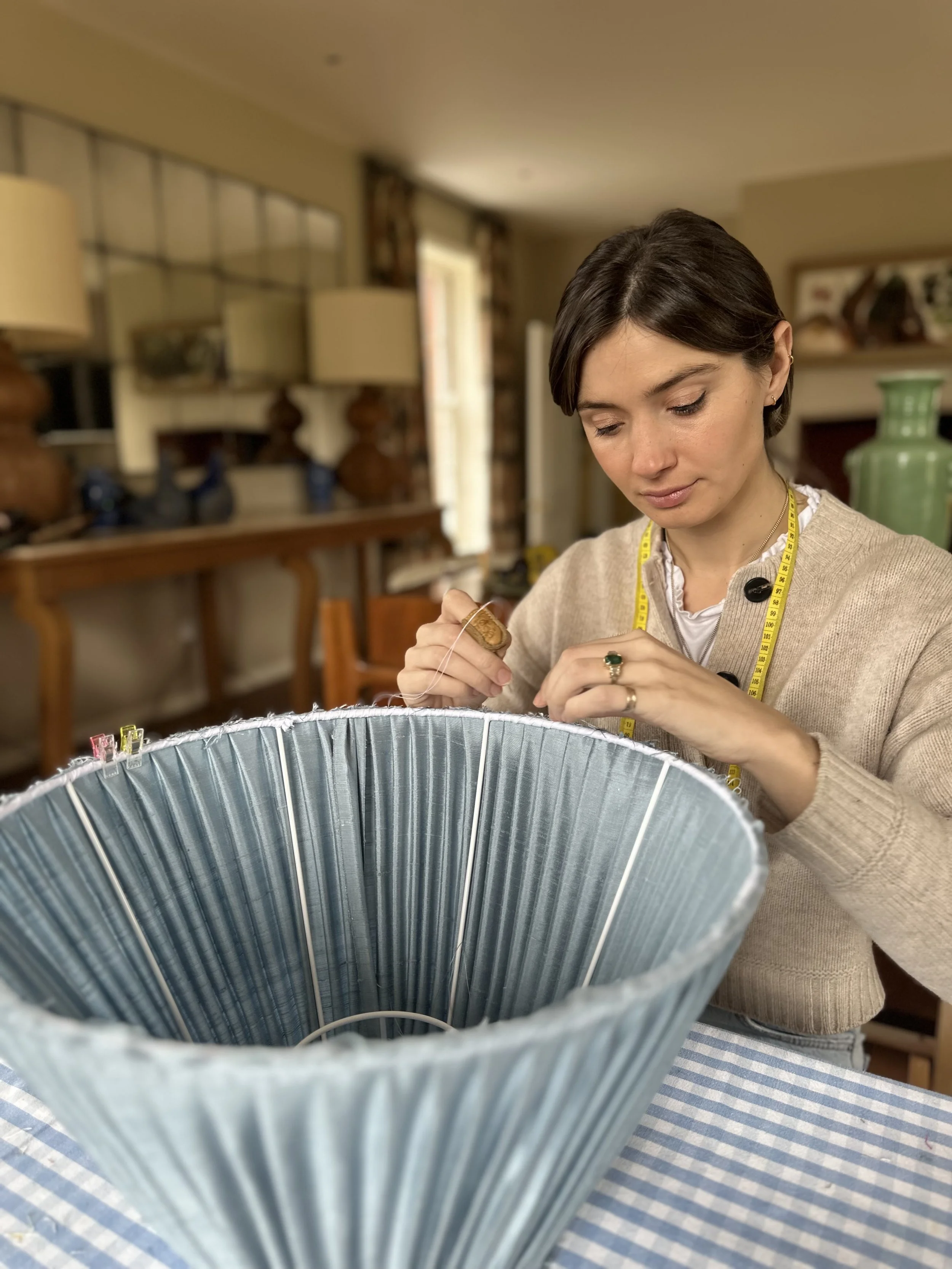 Woman sewing with a tape measure around her neck, working on a lampshade in a cozy home setting.