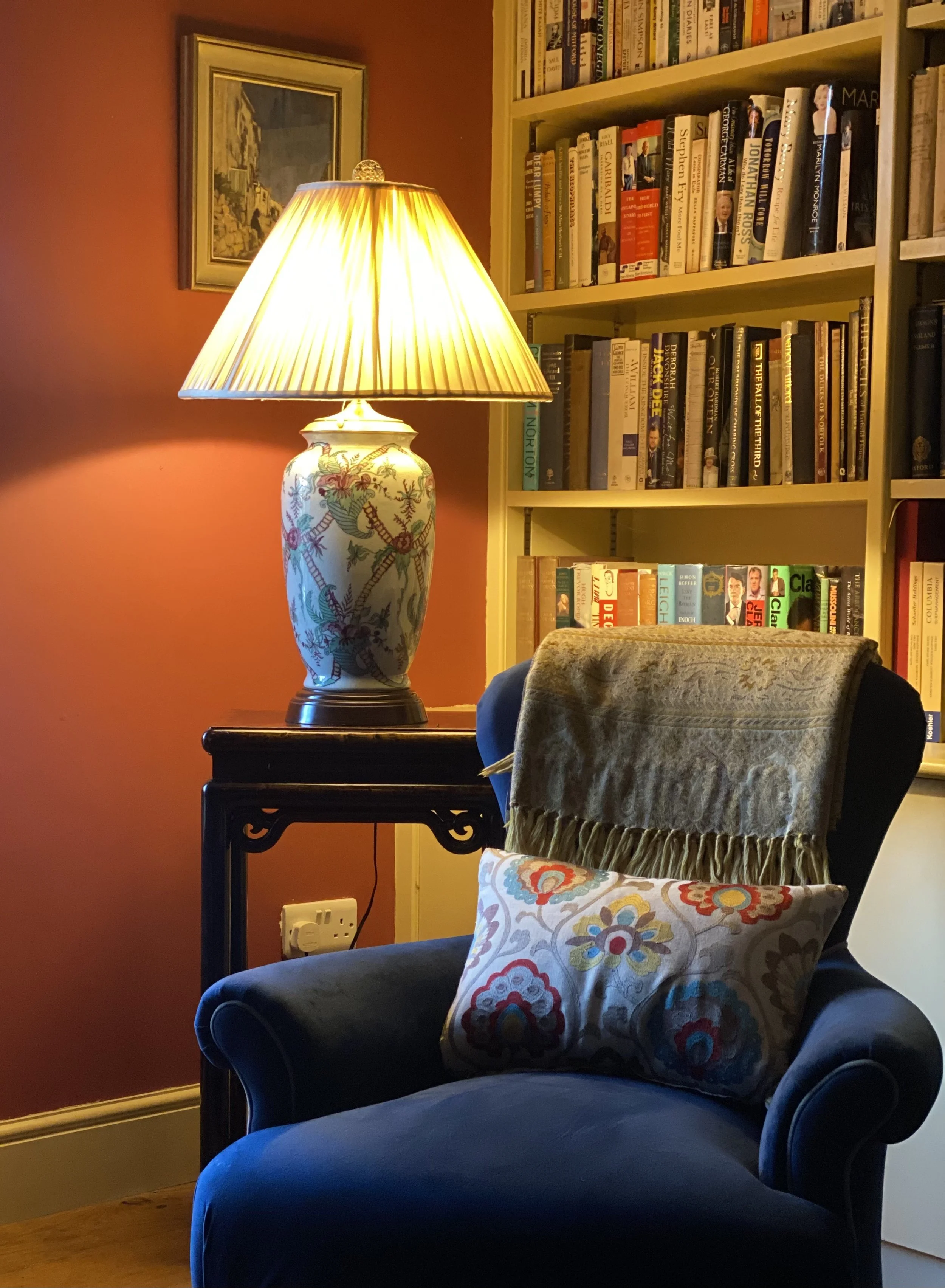 A cozy reading corner with a dark blue armchair, a decorative pillow with floral embroidery, a beige throw blanket, a small dark wooden side table, and a lamp with a floral patterned base and pleated shade. A yellow bookshelf filled with books is beh