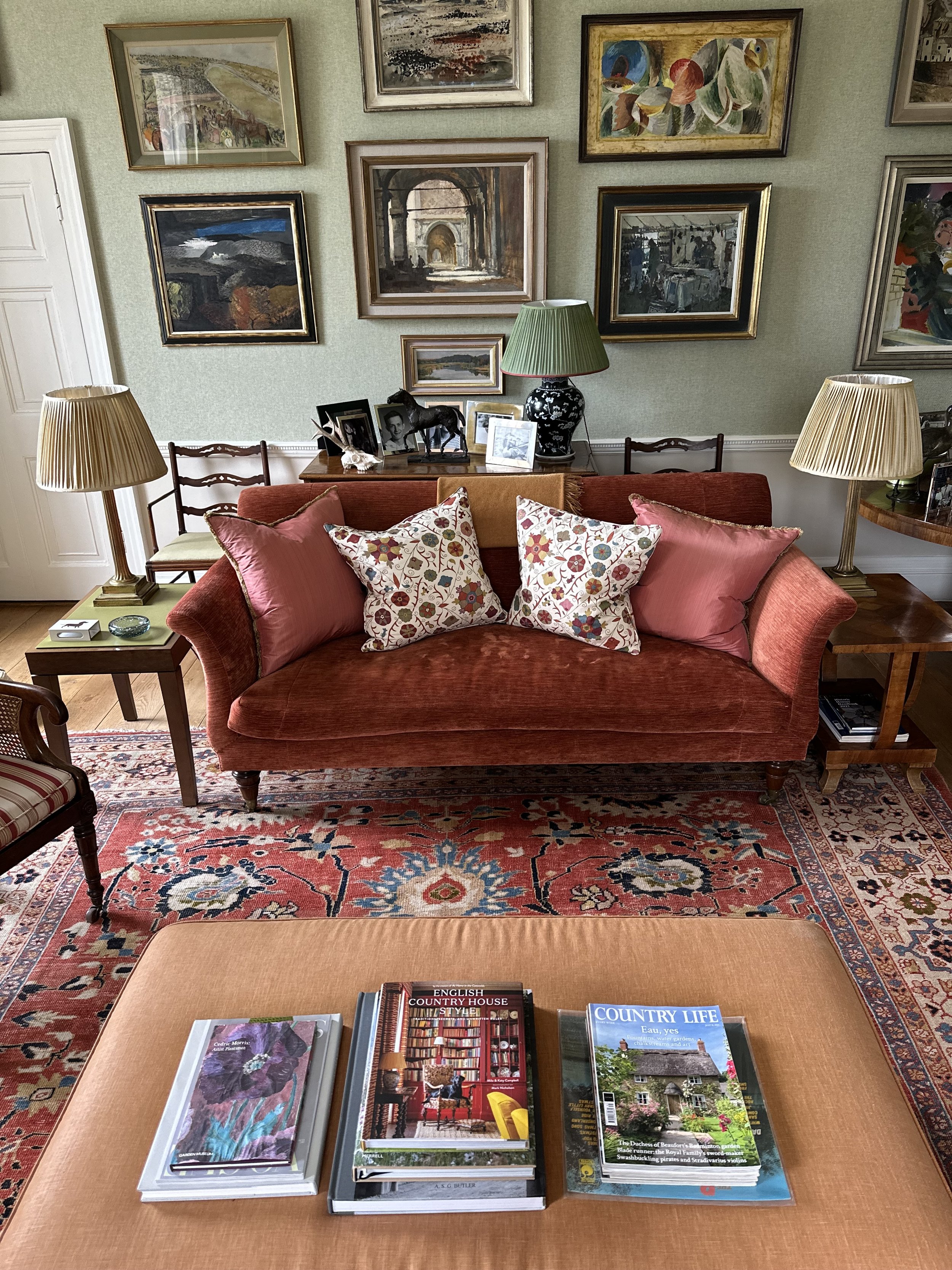A cozy living room with a red velvet sofa holding floral pillows, a wooden side table with lamps on each side, a collection of framed artwork on the wall, and a coffee table in front with three magazines about country houses and country life.