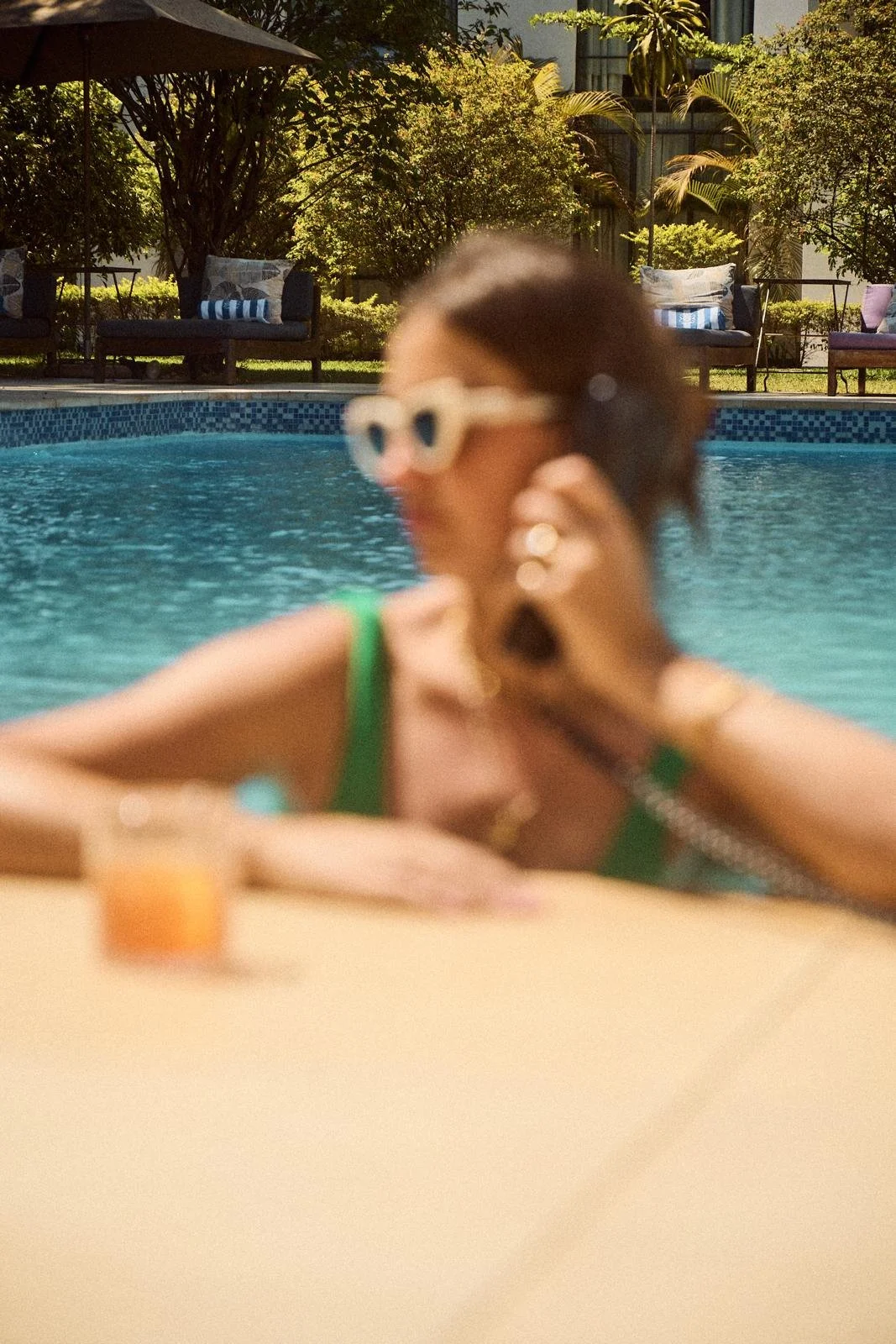 A woman wearing sunglasses and a green swimsuit sitting by a pool, talking on her phone, with trees and outdoor furniture in the background.