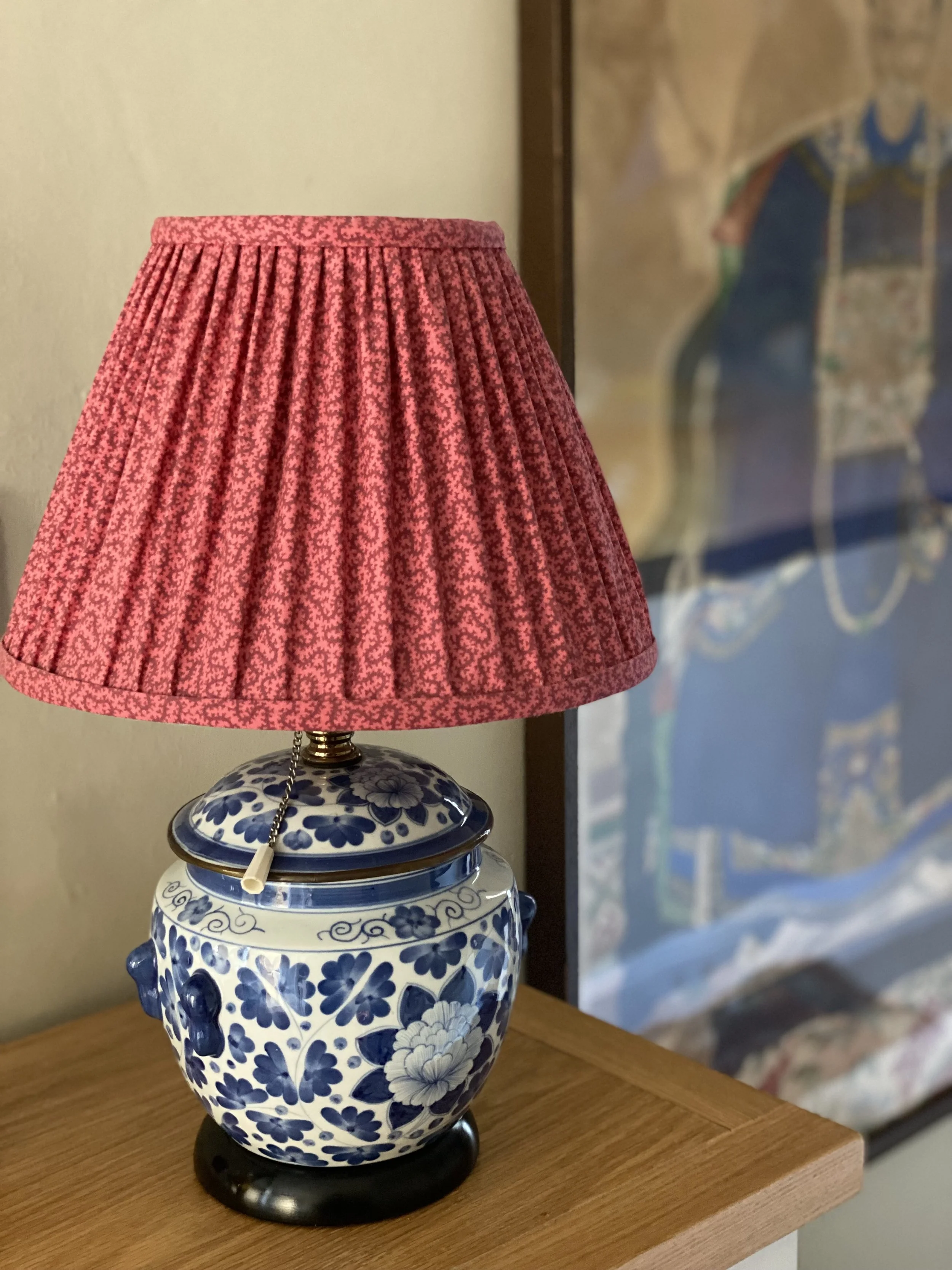 A ceramic table lamp with a blue and white floral pattern, topped with a pink pleated lampshade, placed on a wooden surface.