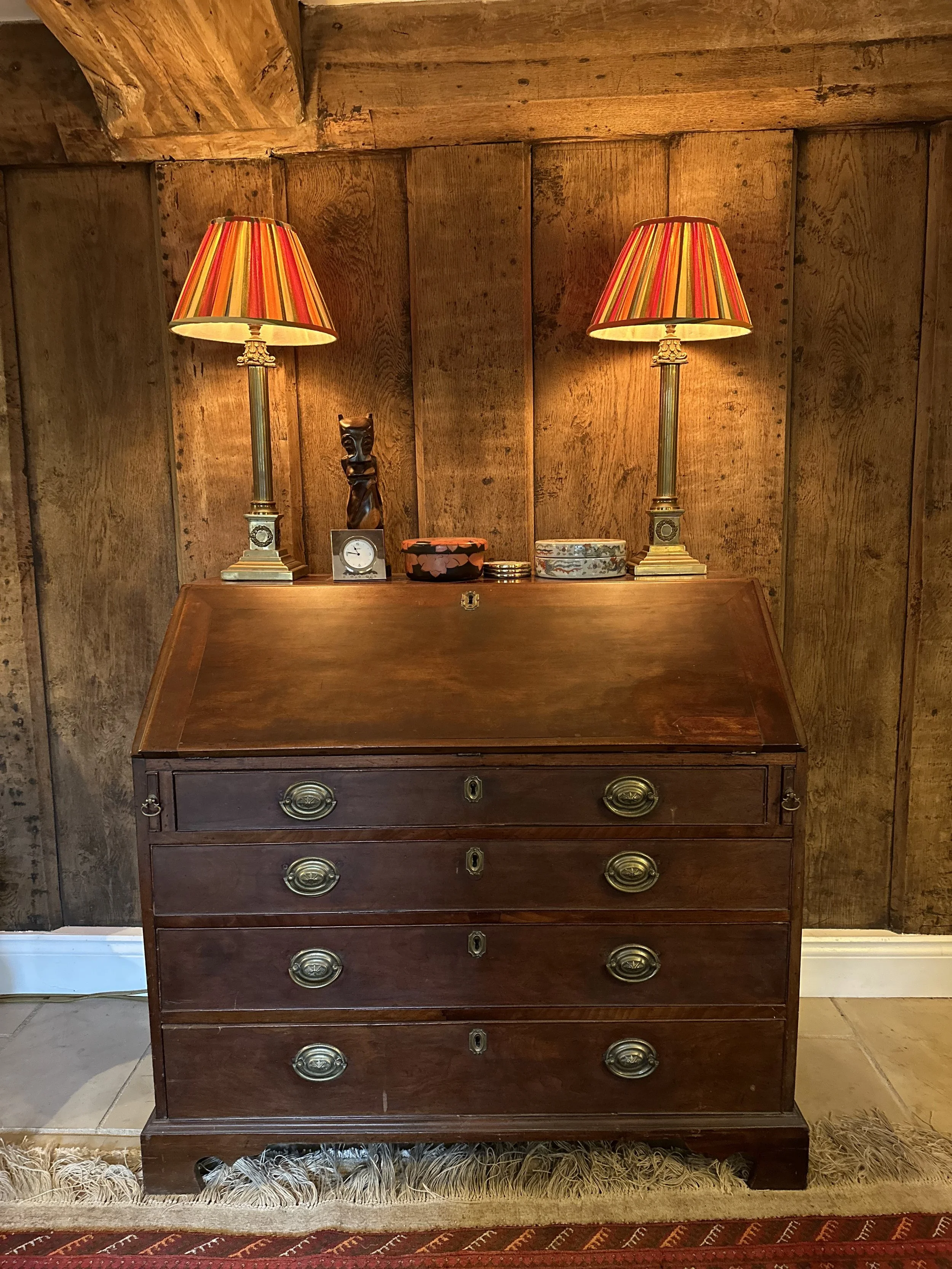 A vintage wooden writing desk with brass handles, topped with two table lamps with striped red, orange, and yellow shades, a small black statue, two boxes, a clock, and some small objects, set against a wooden paneled wall.