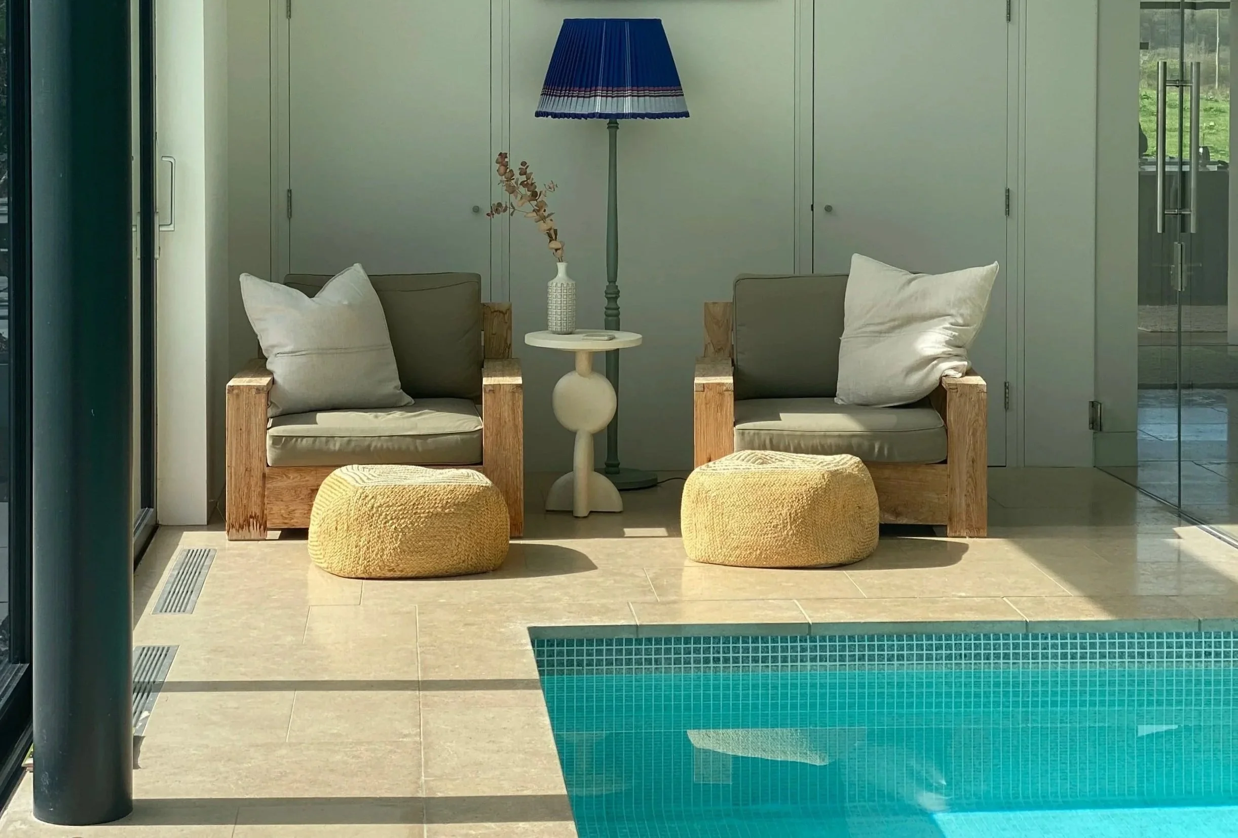 Indoor seating area with two wooden armchairs with gray cushions, a small white side table, a tall floor lamp with a blue lampshade, and two yellow poufs on a beige tiled floor, near a swimming pool with turquoise water.