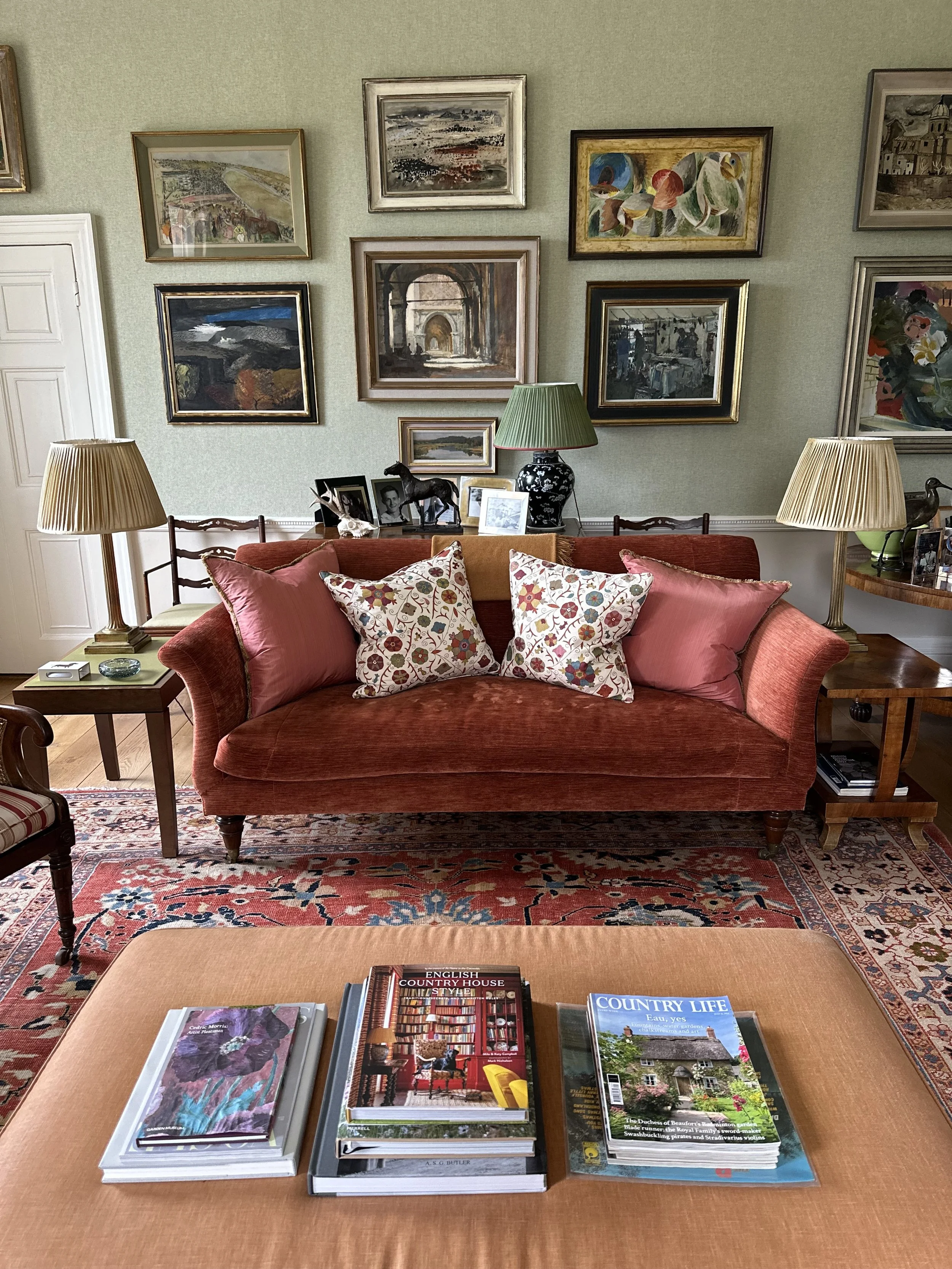 A cozy living room with a red velvet sofa, pink and floral cushions, and a rug. There are framed paintings on the wall, two table lamps, and a stack of magazines on a low table in the foreground.
