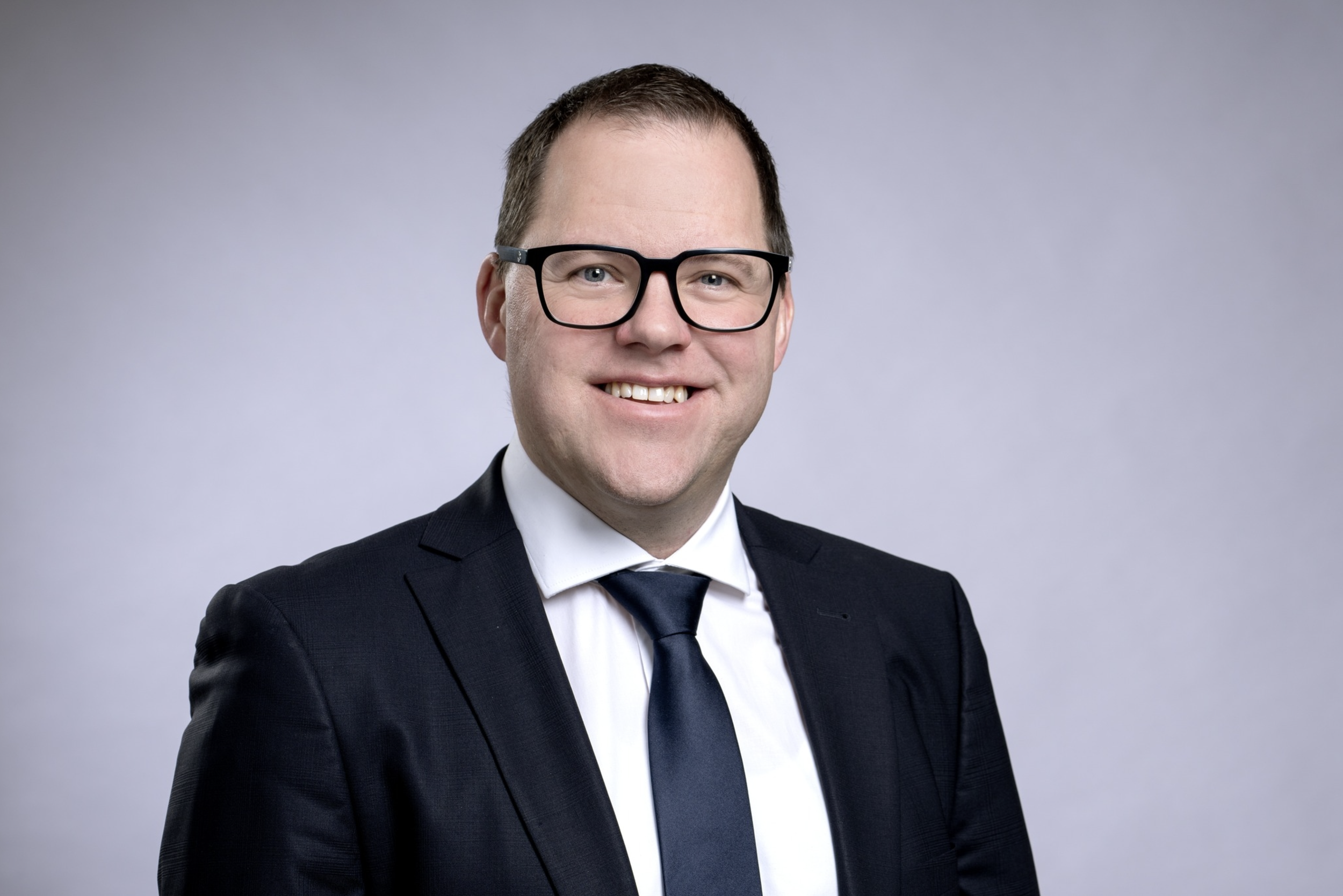 Ein Mann in formeller Kleidung, schwarz-weißer Anzug, Krawatte, trägt schwarze Brille, lächelt freundlich, vor hellem grauem Hintergrund.
