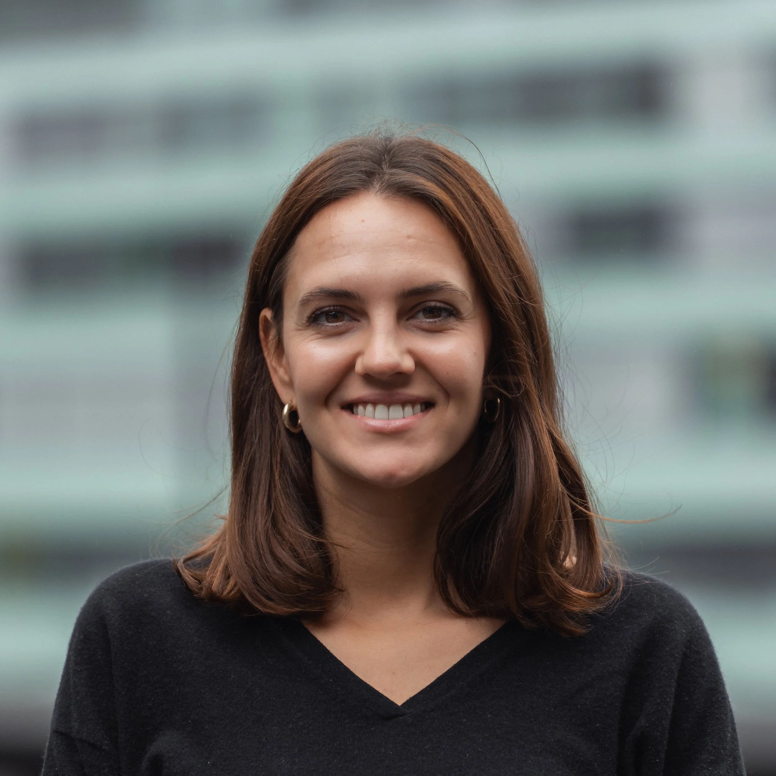 Nahaufnahme des Oberkörpers und Gesichts einer lächelnden Frau mit braunen Haaren, schwarzen Top, in einem unscharfen grauen und grünen Hintergrund.