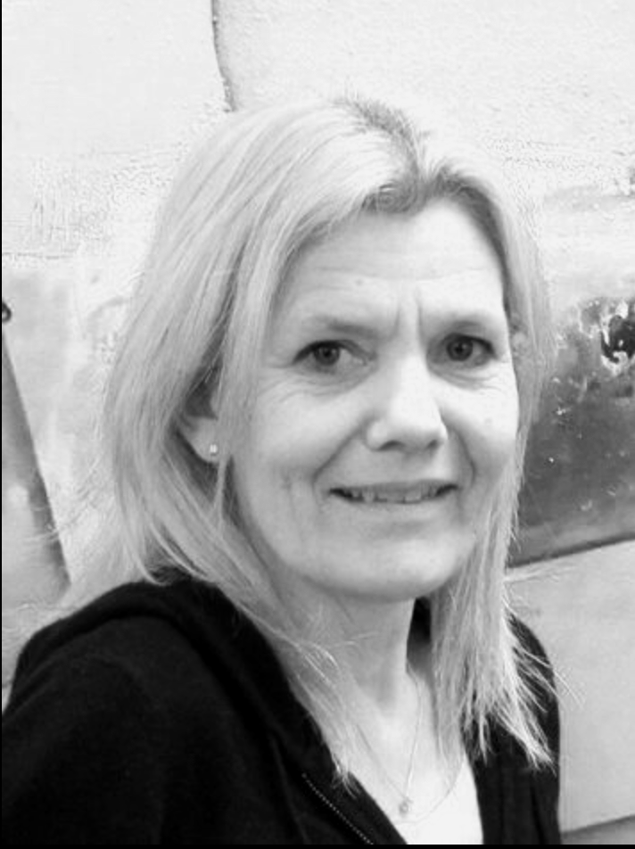 Schwarz-Weiß-Foto einer lachenden Frau mit blondem, schulterlangem Haar, die eine schwarze Jacke trägt, vor einer Wand mit einer künstlerischen Wandmalerei.