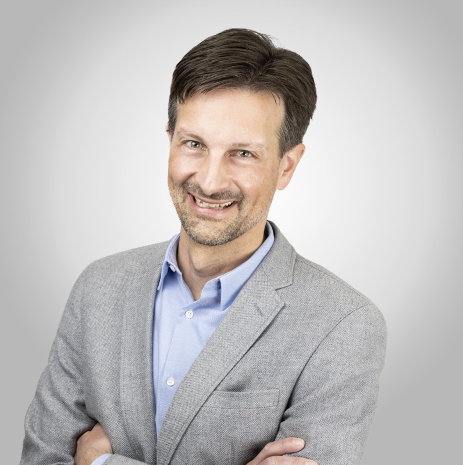 Porträt eines lächelnden, bunten Mannes mit braunen Haaren, blauen Augen, grauem Blazer und blauer Hemd vor grauem Hintergrund.