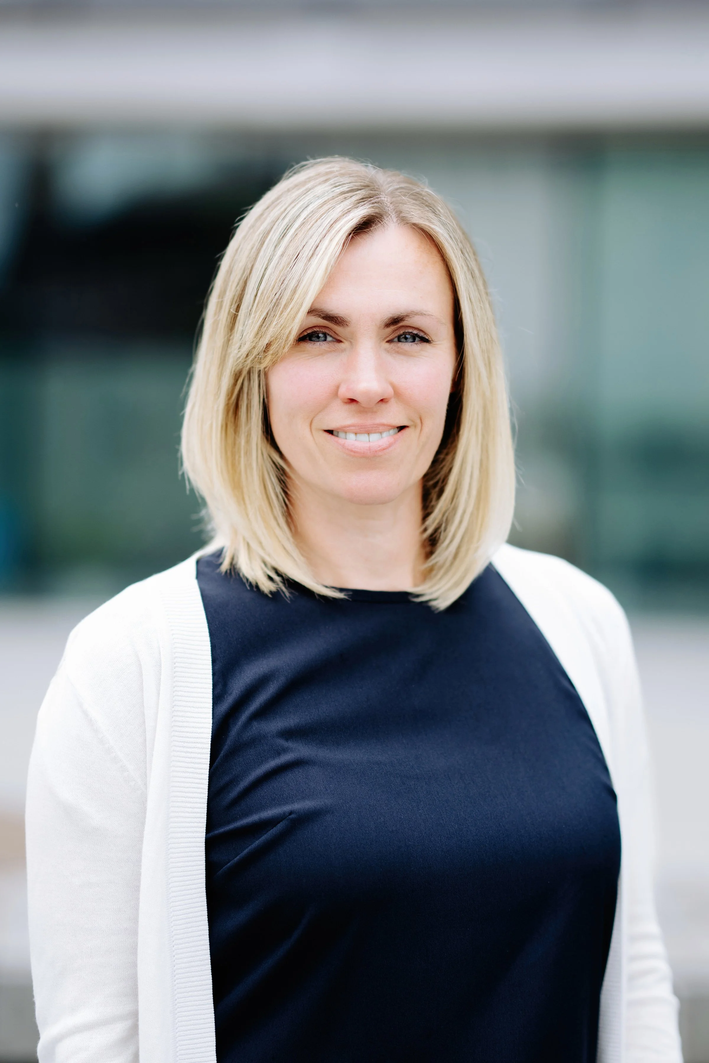 Claire Abbs photo. A blonde woman in a dark top and white cardigan smiling outdoors.