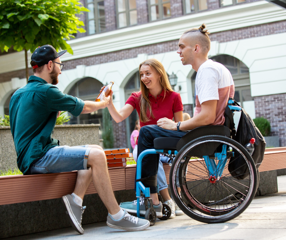 A young woman in a red shirt giving a handshake to a man selling a drink. A man in a wheelchair sits beside her, smiling. They are outdoors near a building with arched windows and trees.