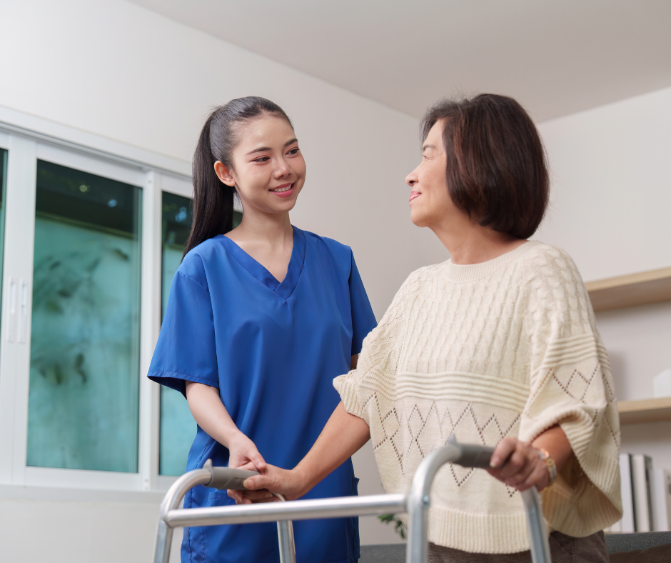 A young female healthcare professional in blue scrubs smiling and holding hands of an older woman using a walker in a bright room.