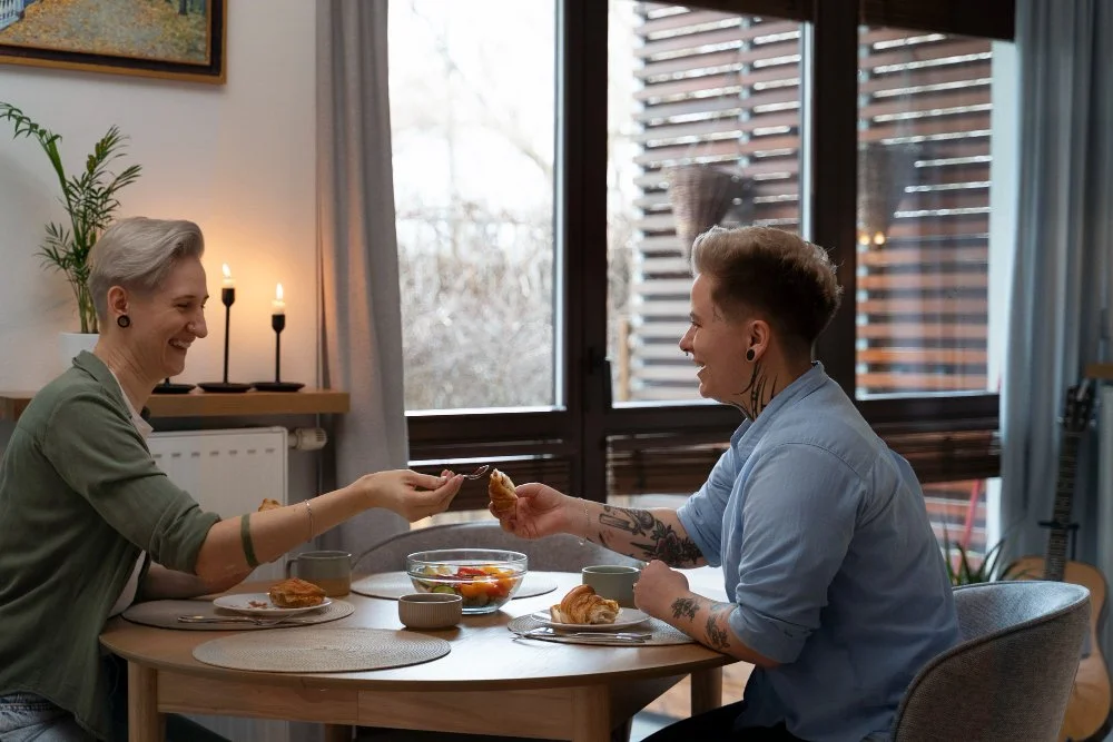 Two women with short gray and brown hair sitting at a round wooden table, sharing breakfast in a bright, cozy room with large windows and wooden slat blinds. They are smiling, passing a piece of croissant, with a bowl of fruit and coffee mugs on the table.