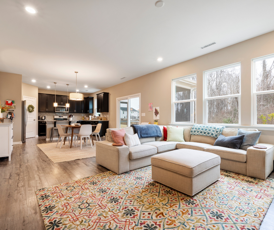 Bright living room with beige walls, large windows, and a light-colored sectional sofa with various pillows. Adjacent is a dining area with a round table and six chairs, and a modern kitchen with dark cabinets and pendant lighting.